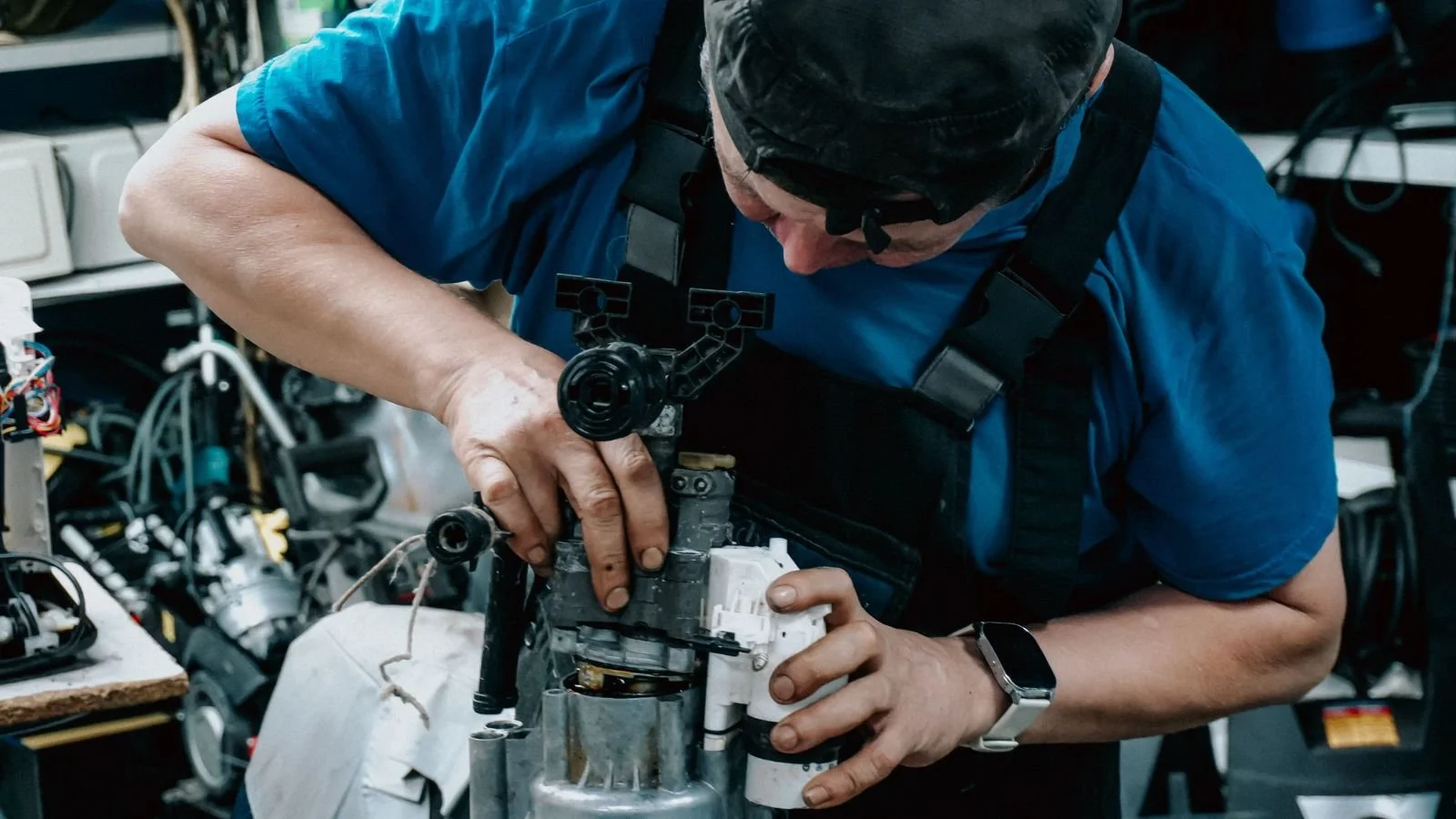 A person working on a mechanical device with various wires and components around, wearing a blue shirt, black cap, and a watch.