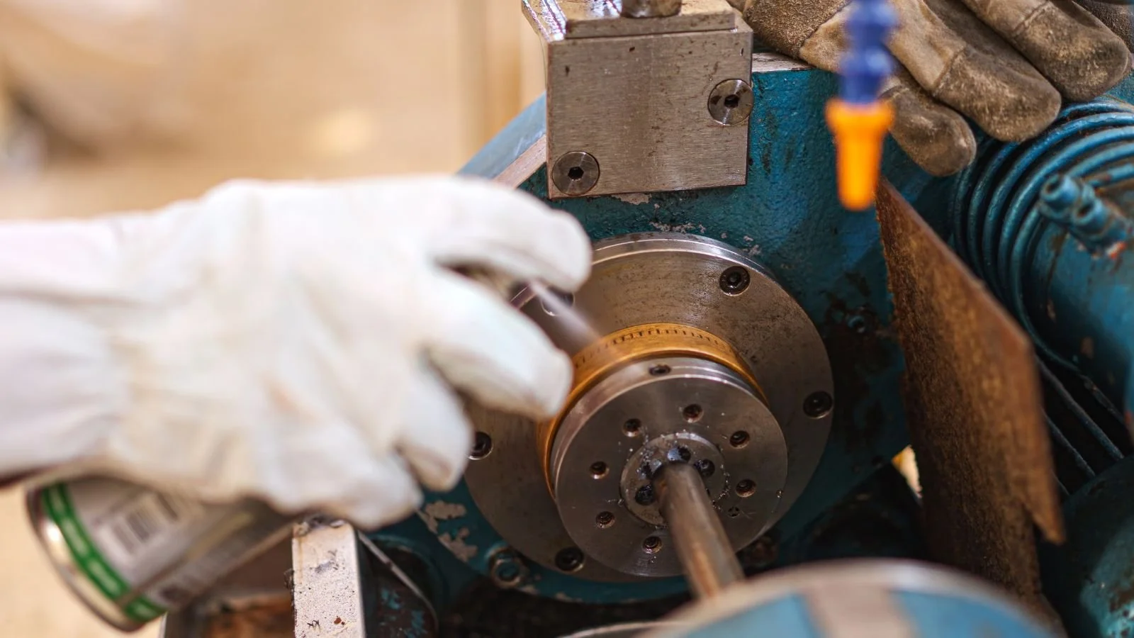 Close-up of a person wearing white gloves working on a mechanical component using a metal tool, with a blue machine and some wiring nearby.