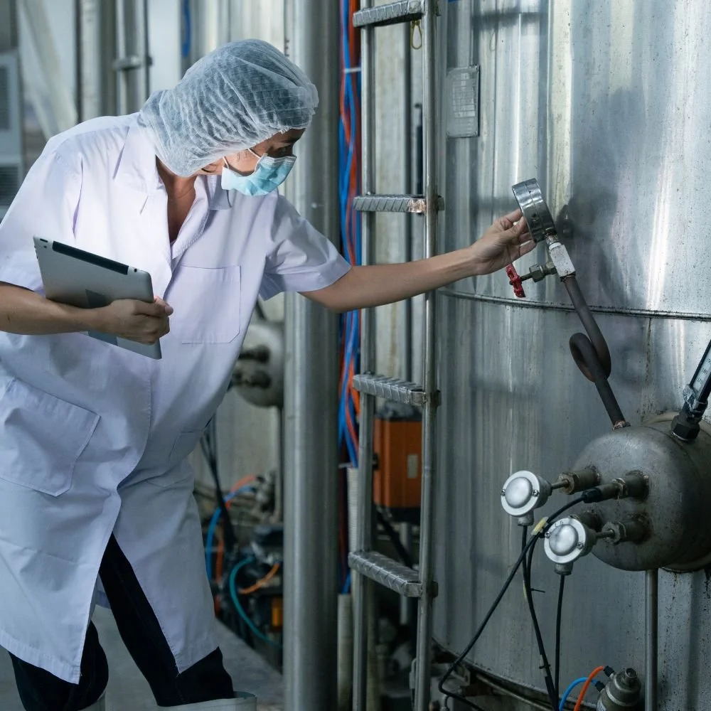 A scientist or engineer in protective gear checking equipment on a stainless steel industrial tank in a laboratory or manufacturing setting.