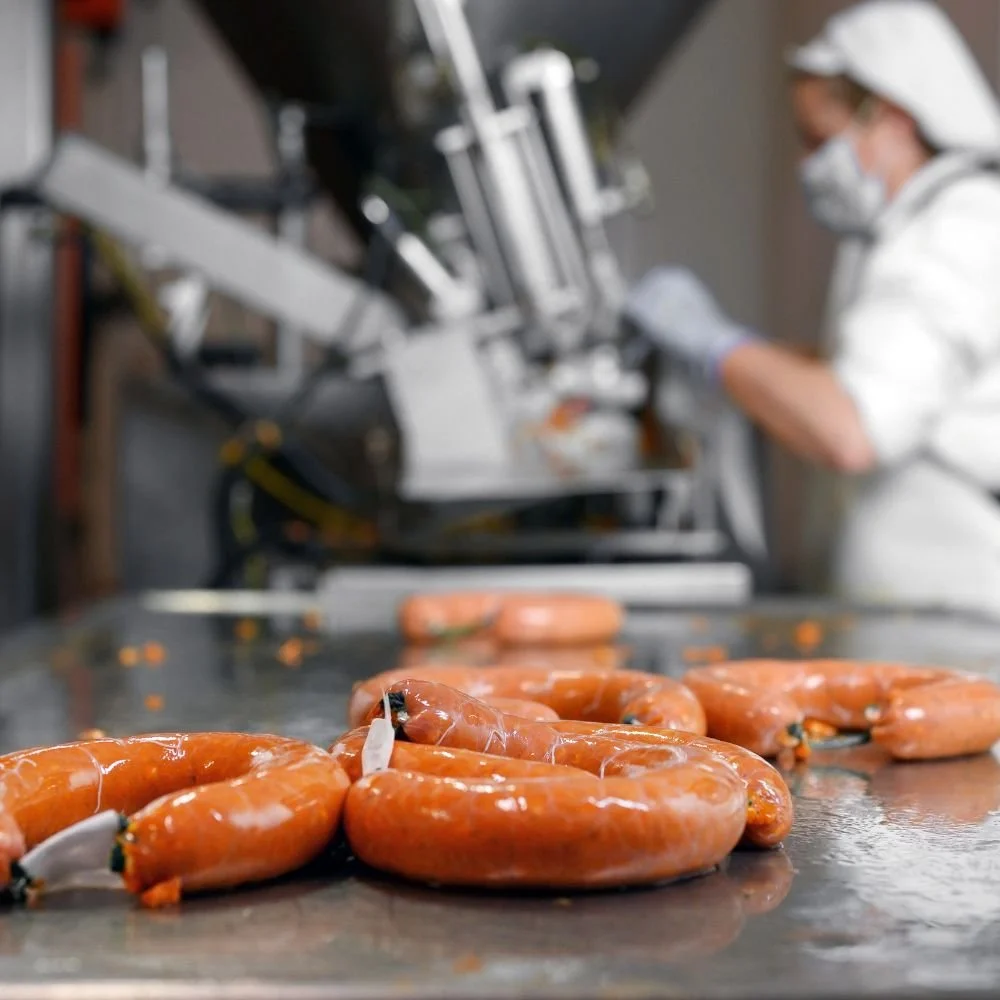 Close-up of sausages on a butcher's counter with a worker in protective clothing and a mask working in the background.