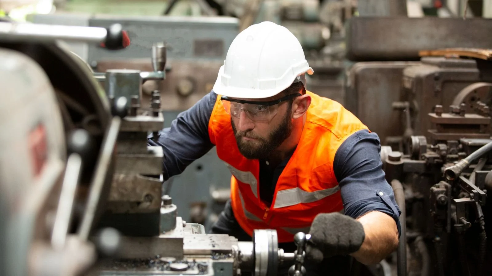 A male factory worker wearing a white safety helmet, orange safety vest, glasses, and gloves operating machinery in an industrial setting.