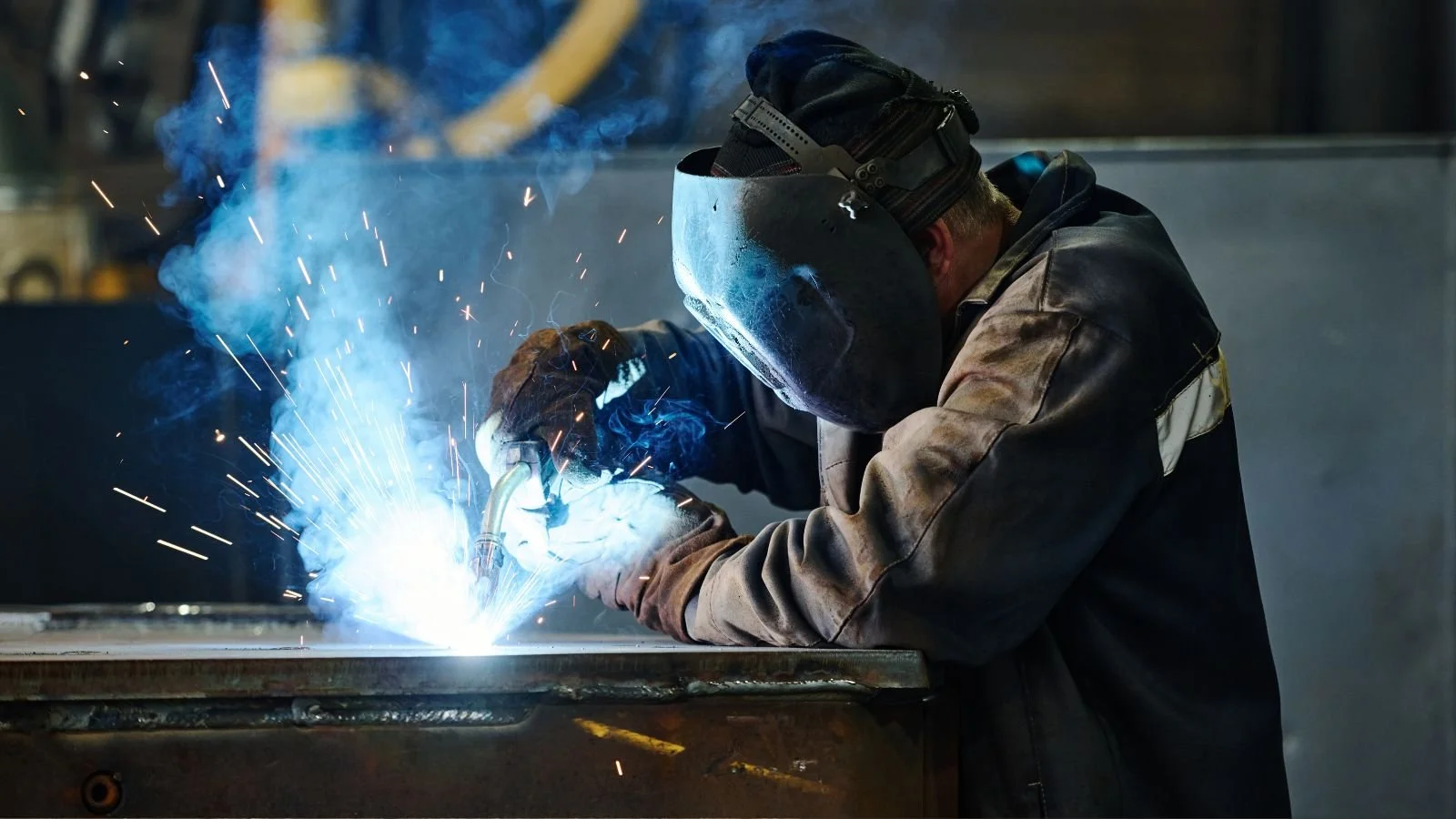 A worker welding metal in a factory, wearing protective gear and a welding helmet, with sparks and blue light from the welding process.