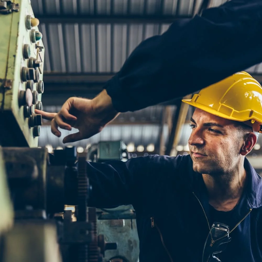 A man wearing a yellow safety helmet operating industrial machinery with the assistance of another person in a dark uniform.