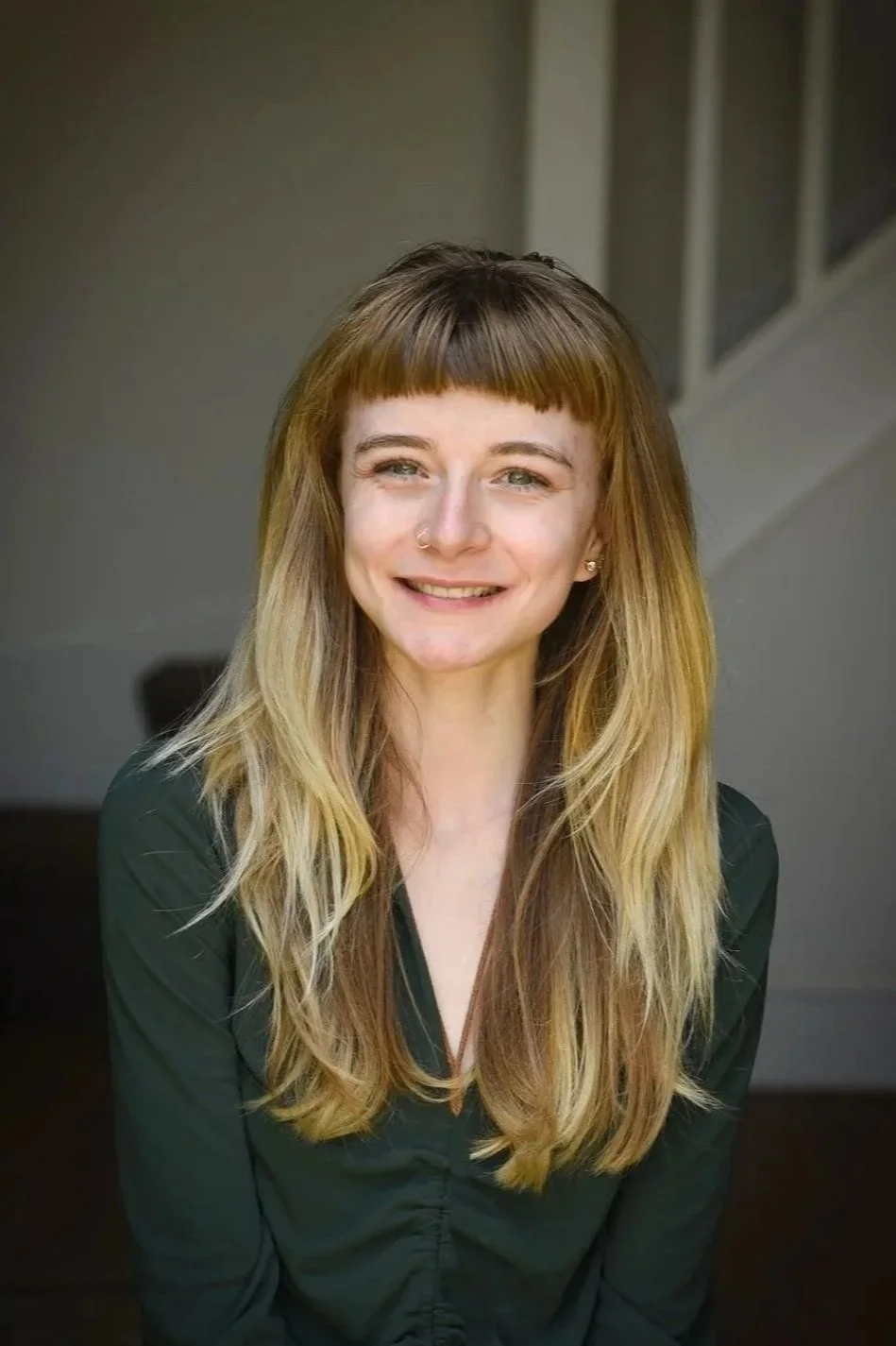 A woman with long, wavy blonde hair and a nose ring smiling indoors.