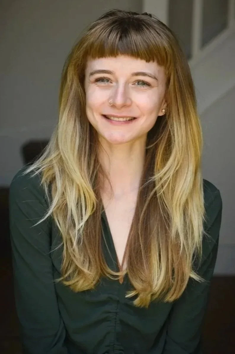 Young woman with shoulder-length, wavy blonde hair and bangs, smiling at the camera, wearing a dark green top, indoors.