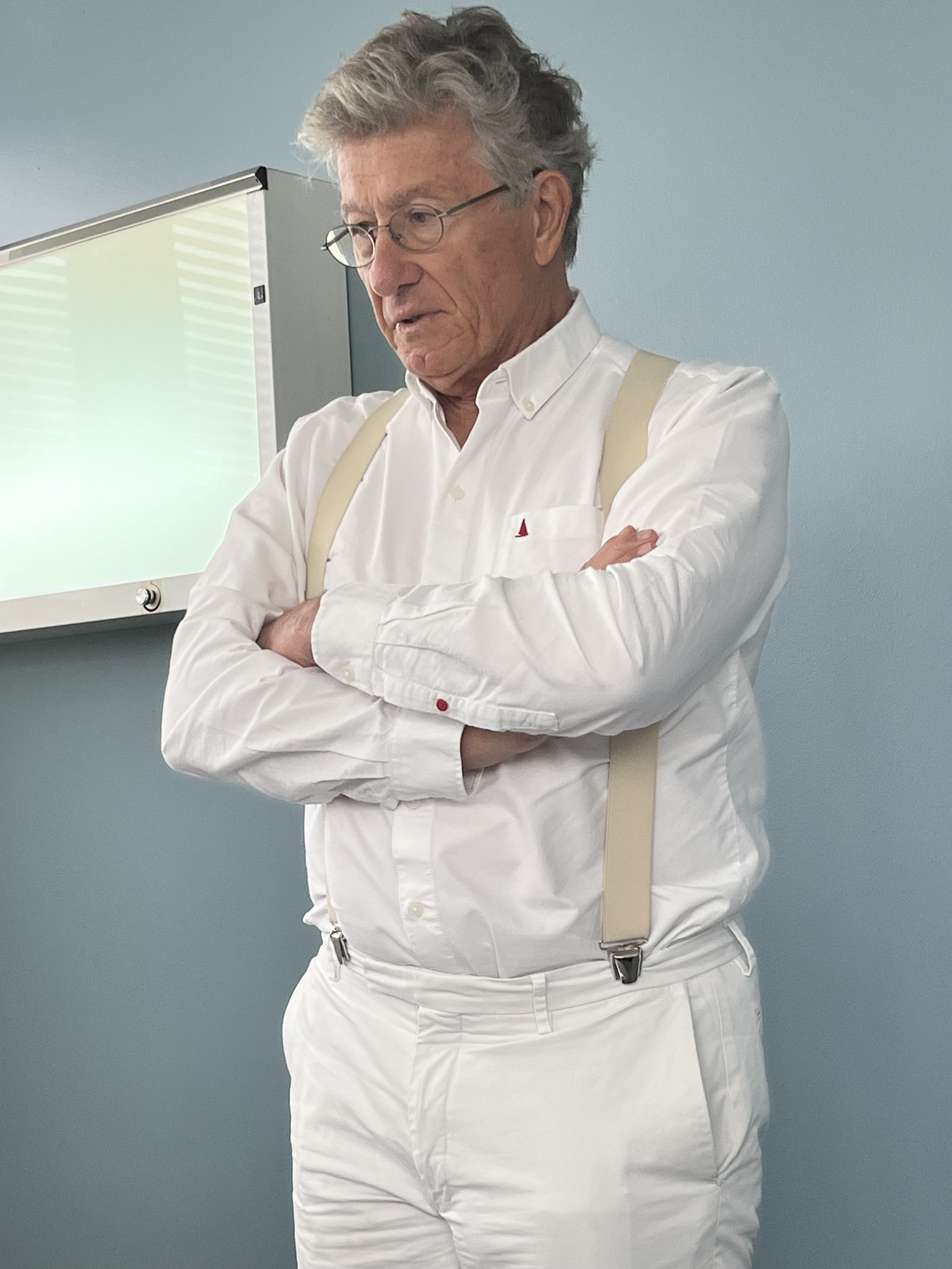 Un homme âgé habillé en chemise blanche et pantalon blanc, portant des bretelles, debout contre un mur bleu, avec des lunettes rondes et des cheveux gris, semblant concentré ou pensif.