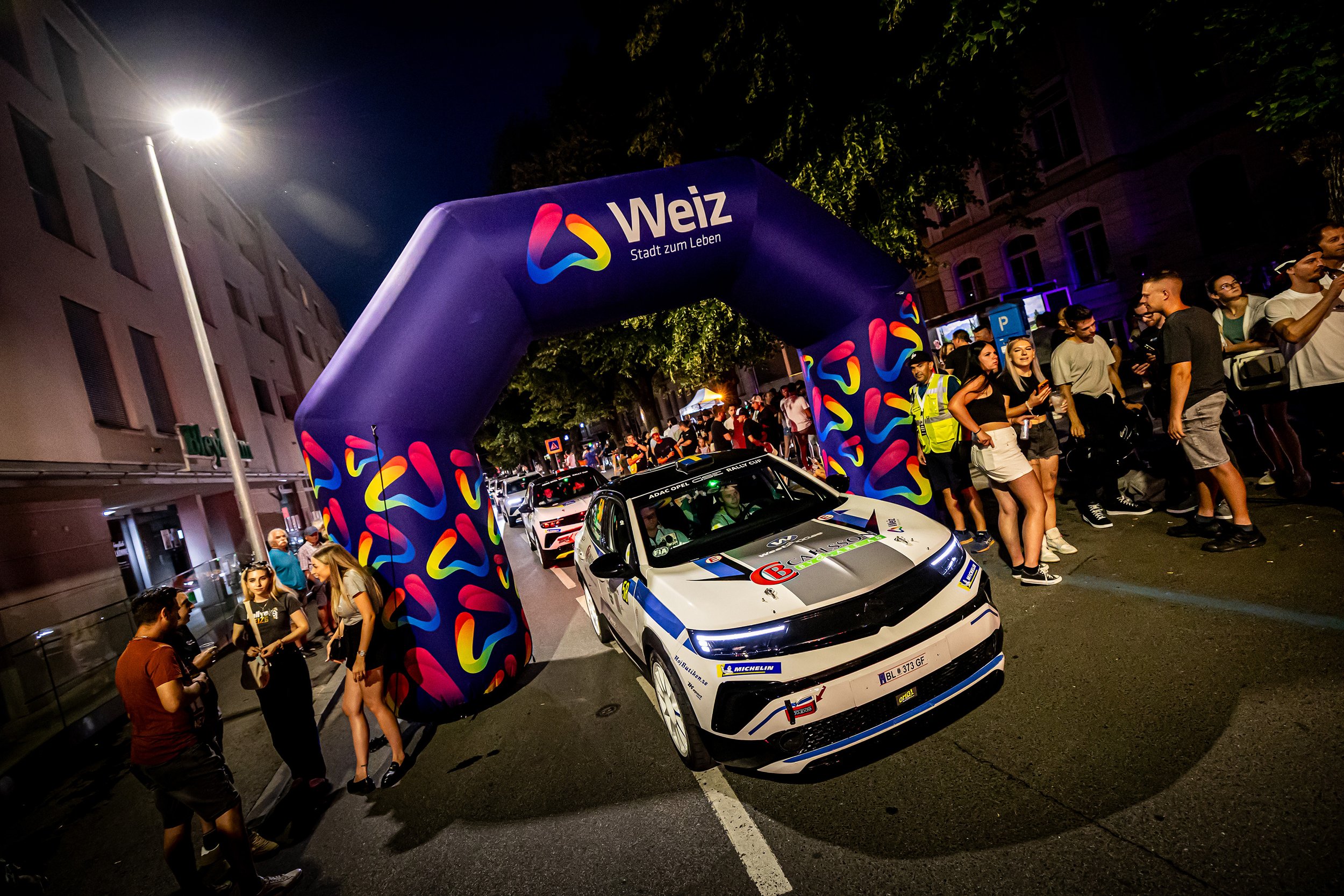 Kvällsbild av ett rallybilstävling i Weiz, Österrike, med publik och bilar på gatan under en upplyst tävling.