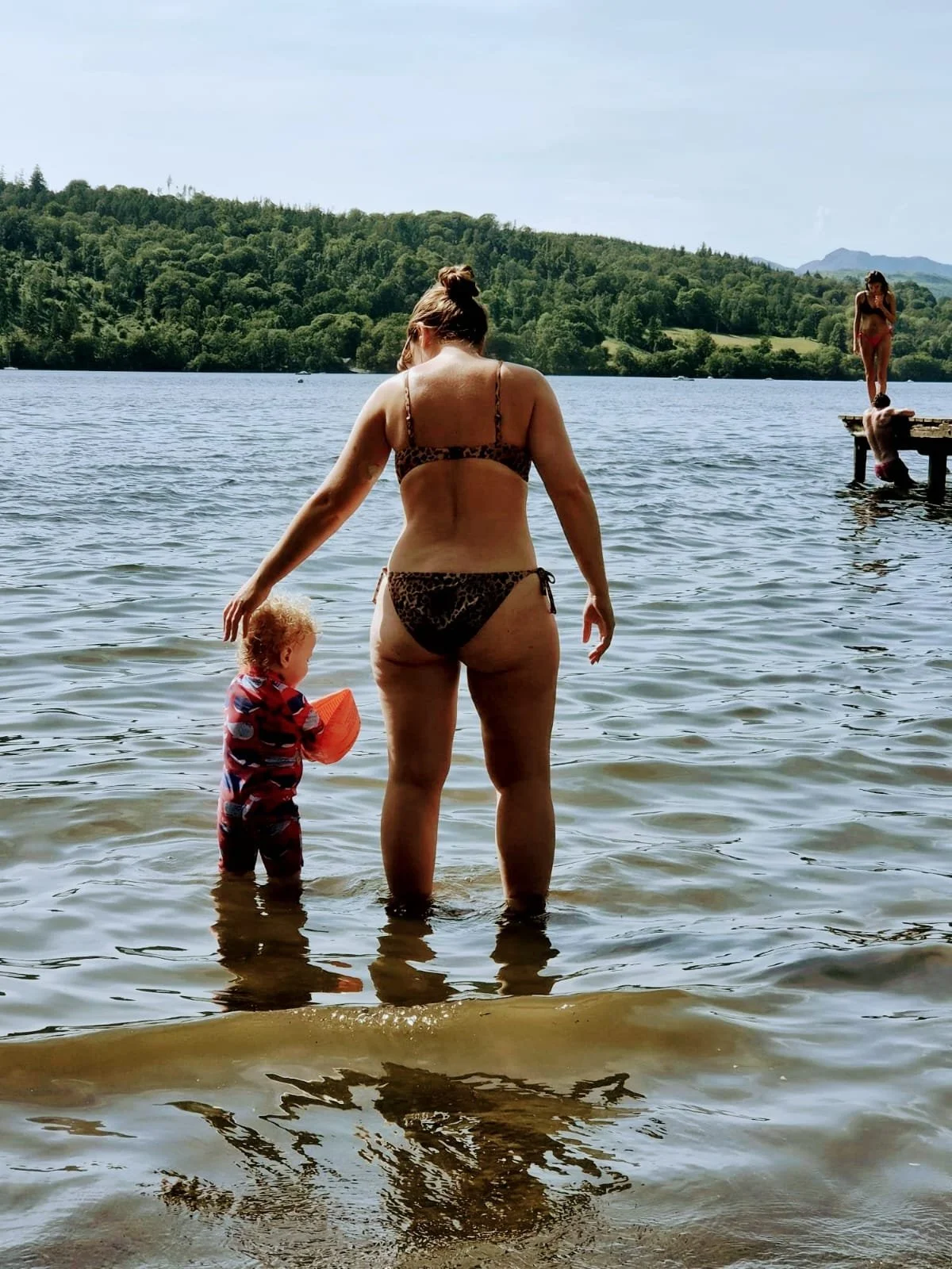 A woman in a leopard print bikini standing in a lake with a young child holding a floaty. In the background, a woman is standing on a dock leaning forward.