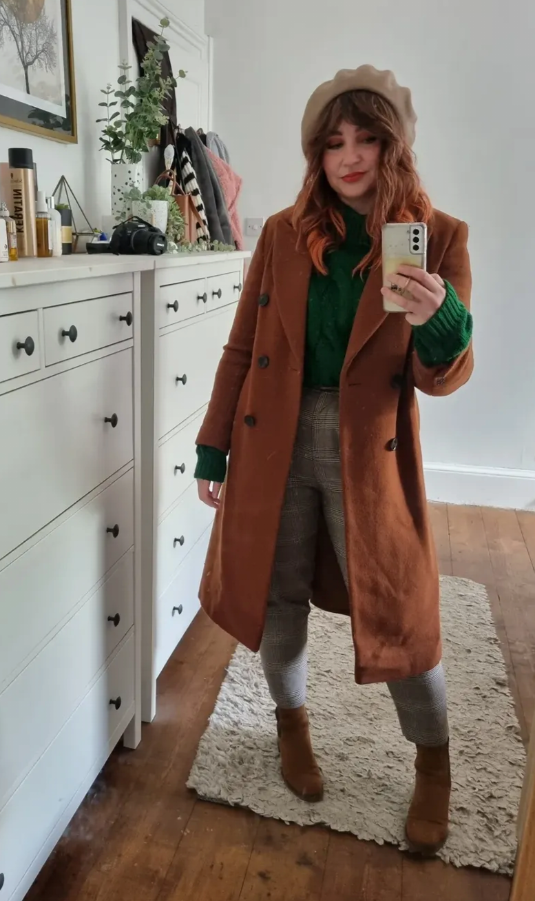 Woman taking a mirror selfie indoors, wearing a beige beret, brown coat, green sweater, plaid pants, and brown ankle boots, standing on a cream-colored rug next to a white dresser with various items and a mirror.