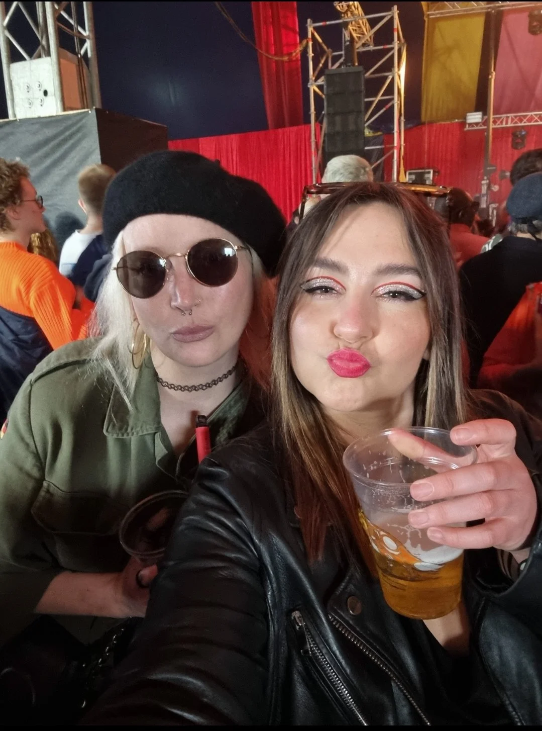 Two women taking a selfie at a crowded event or concert, with stage lighting and scaffolding in the background. One woman has long hair, makeup, and a leather jacket, holding a drink. The other has white hair, sunglasses, and an olive green jacket, also holding a drink.