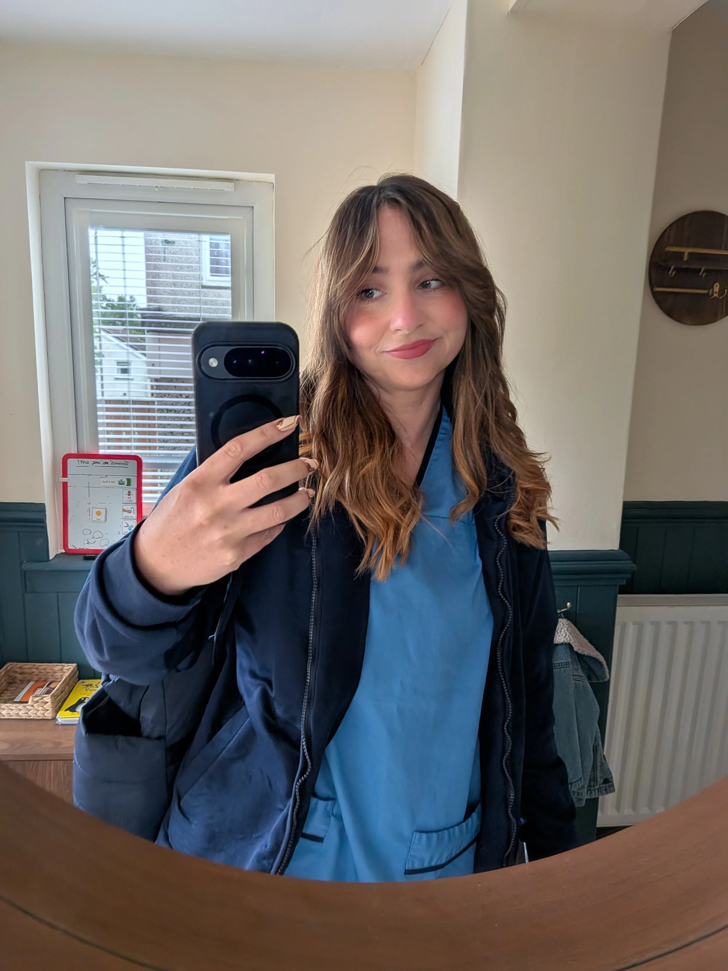 A woman with long, wavy, light brown hair is taking a selfie in a mirror. She is wearing a blue shirt and a dark jacket. The background includes a window with blinds, a radiator, and some household items on a table.