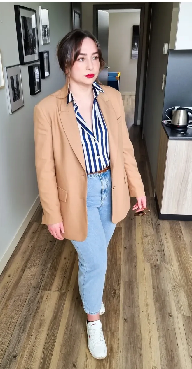 A woman standing indoors on a wooden floor, wearing a tan blazer over a blue and white striped shirt, light blue jeans, and white sneakers, holding sunglasses in her right hand.