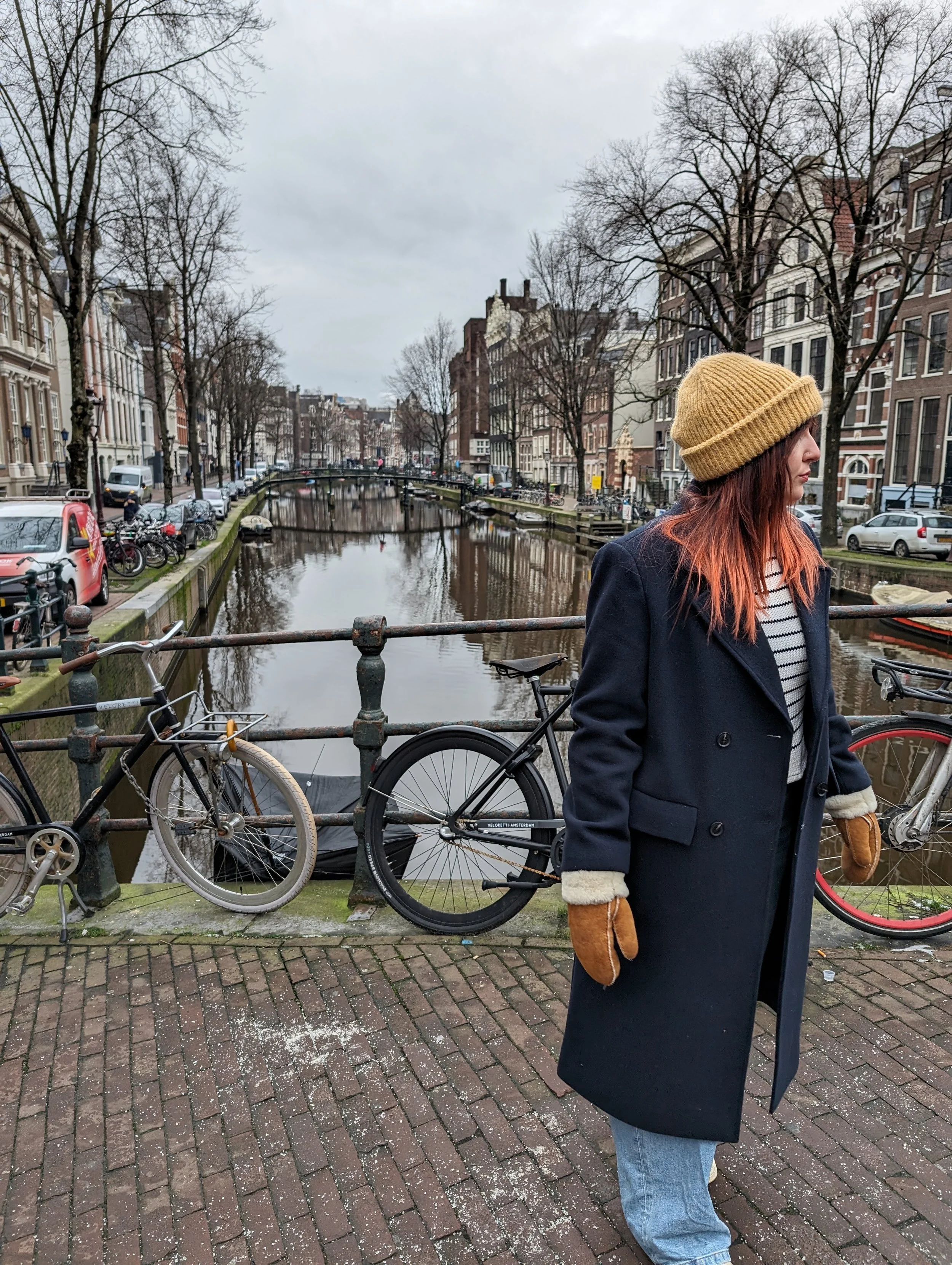 A woman with red hair wearing a tan beanie, navy coat, and brown gloves, standing near bicycles on a brick pathway by a canal in a European city with narrow buildings and leafless trees.
