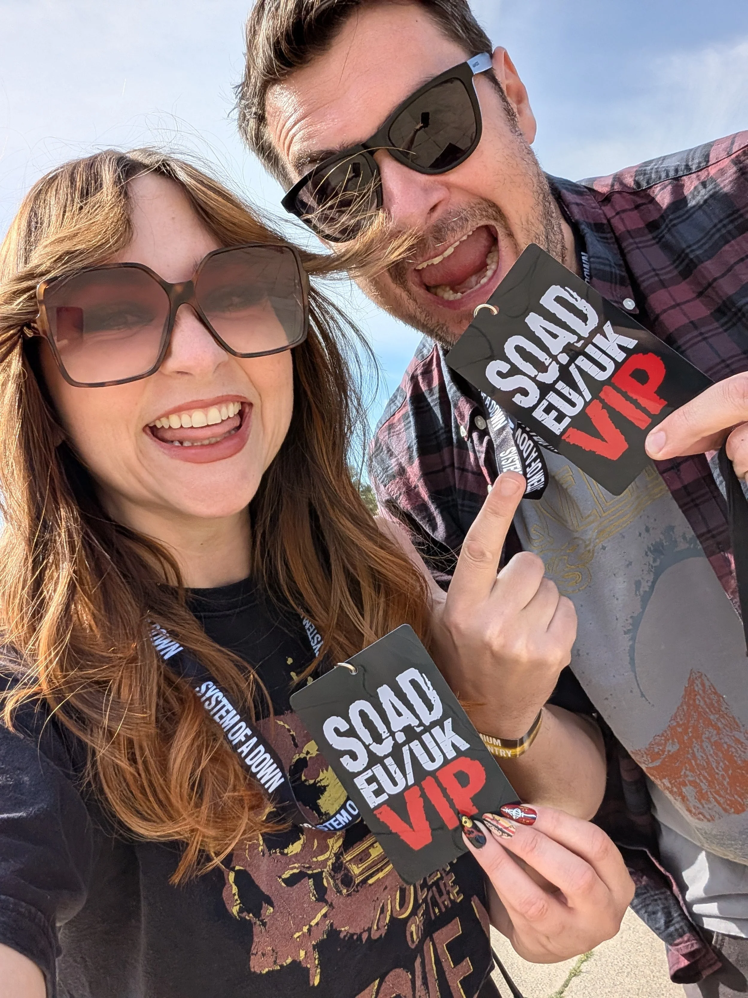 Close-up of two people wearing sunglasses, smiling and holding a sign that says 'Vaccine Shock' with a red 'V'. They are outdoors on a sunny day.