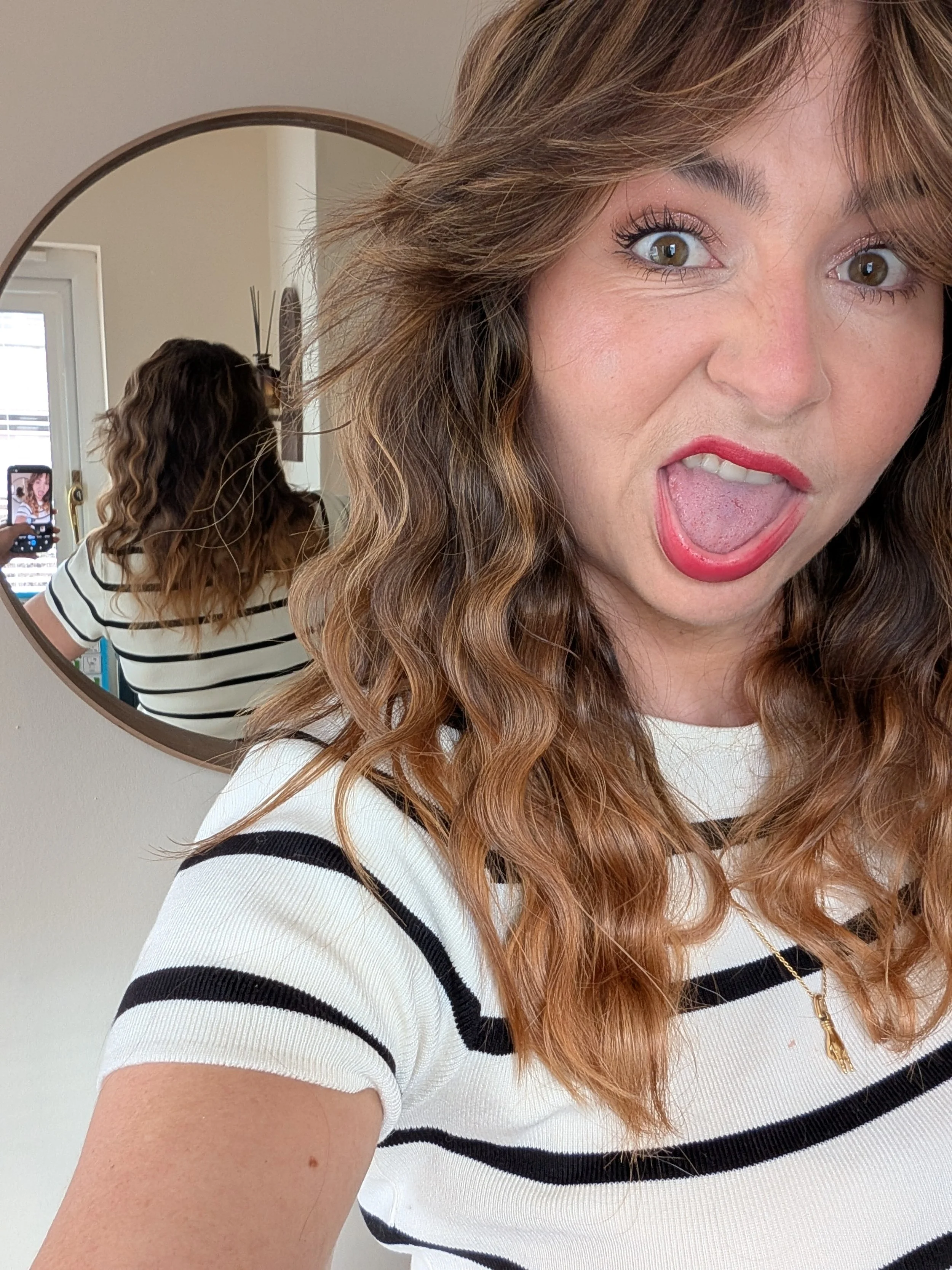 Close-up of a woman with curly brown hair, wearing a white shirt with black horizontal stripes, standing in front of a mirror, taking a selfie with her phone.