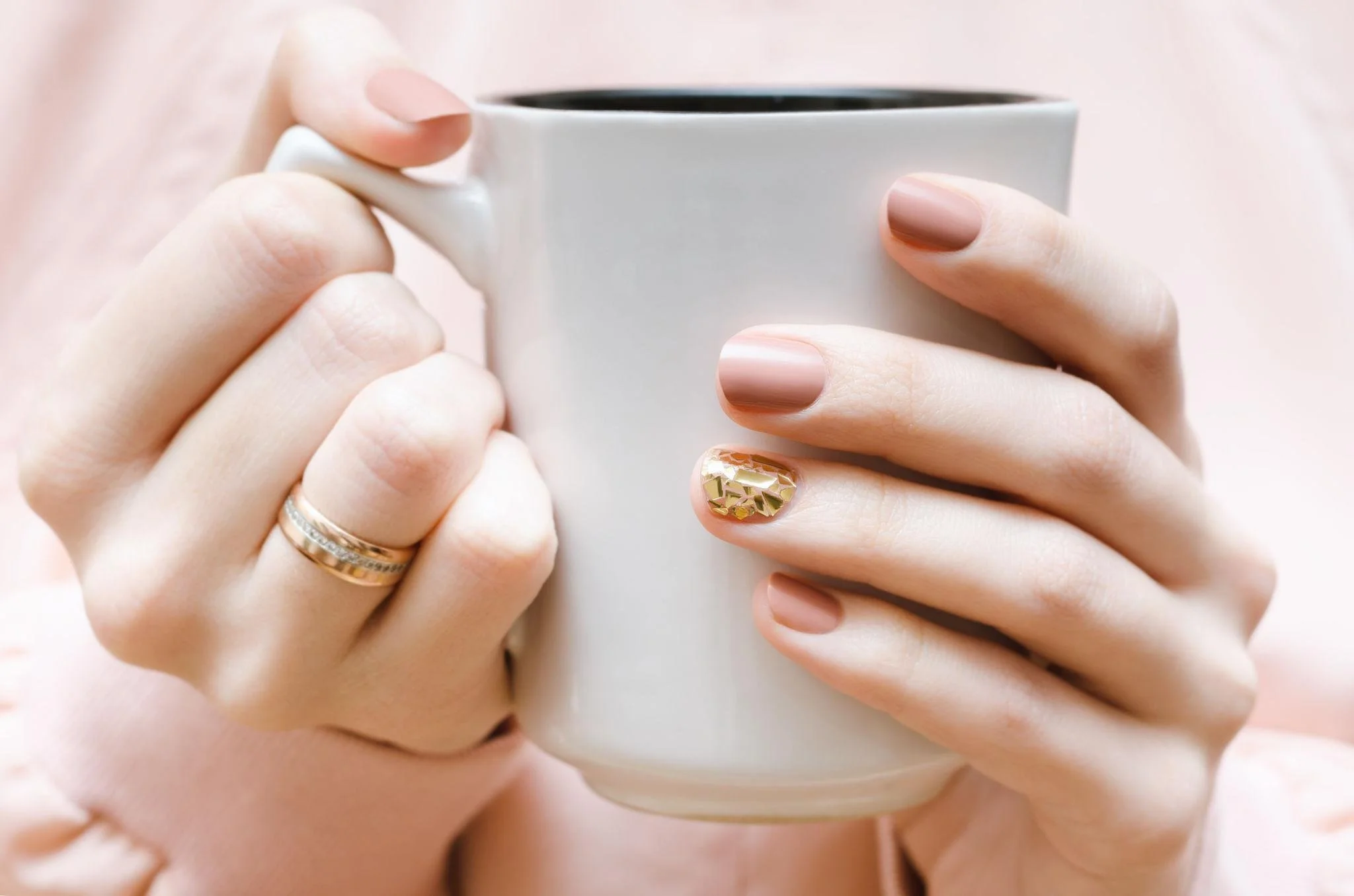 Close-up of hands holding a white mug filled with a dark beverage. The hands have neatly groomed nails, one with metallic gold nail art. The person is wearing multiple rings, and the background is softly blurred.