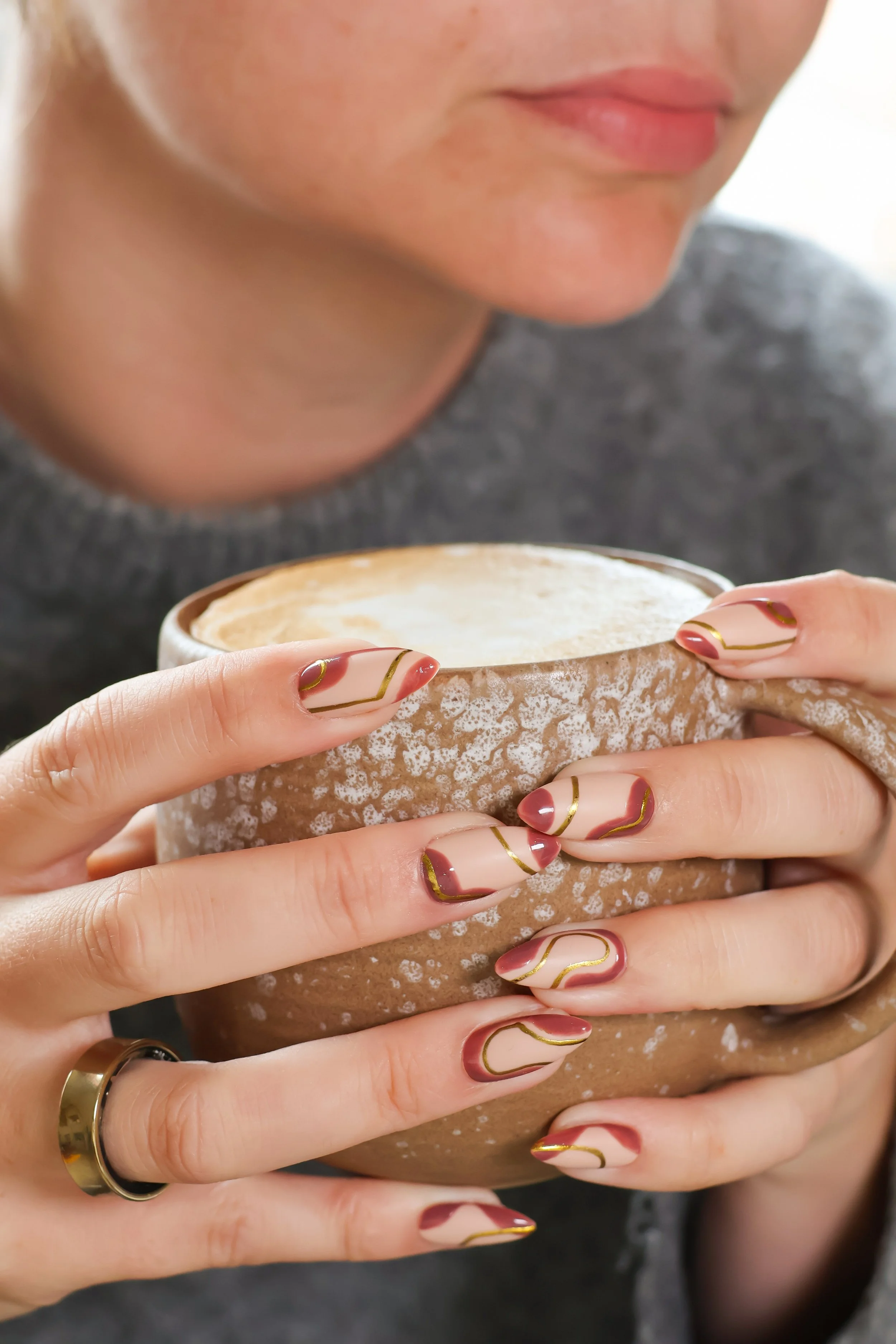 Close-up of a person holding a textured, beige ceramic cup with cream-colored beverage. The person's face is partially visible, showing lips, chin and part of the nose. Their nails are decorated with a modern, abstract design in cream, red and gold. They wear a gold ring on their left index finger. The person is wearing a grey garment.