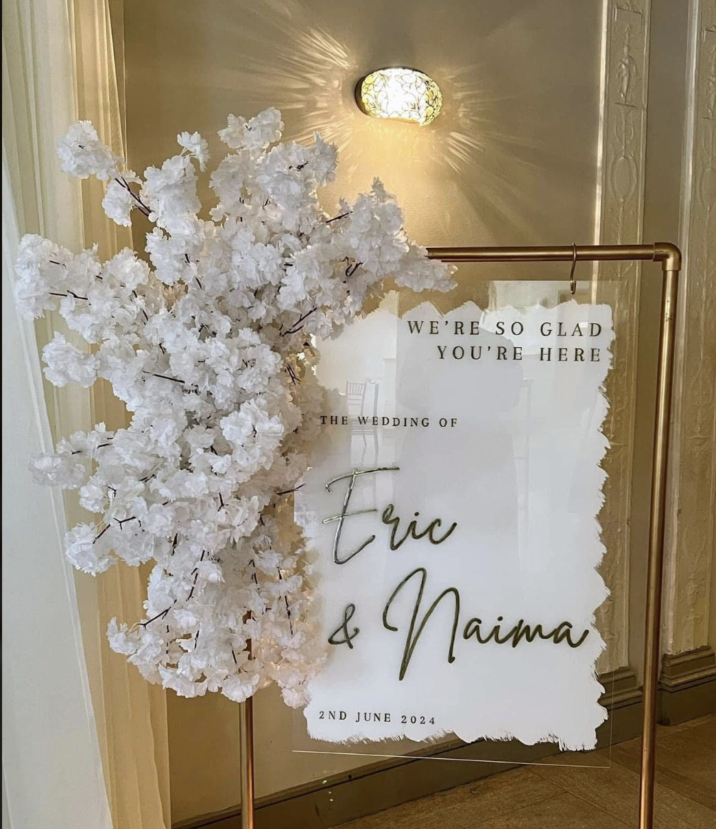 A wedding sign with floral decorations on a stand, displaying the message "We're so glad you're here" and details about the wedding of Erica and Naima on June 2, 2024, in a warm indoor setting.