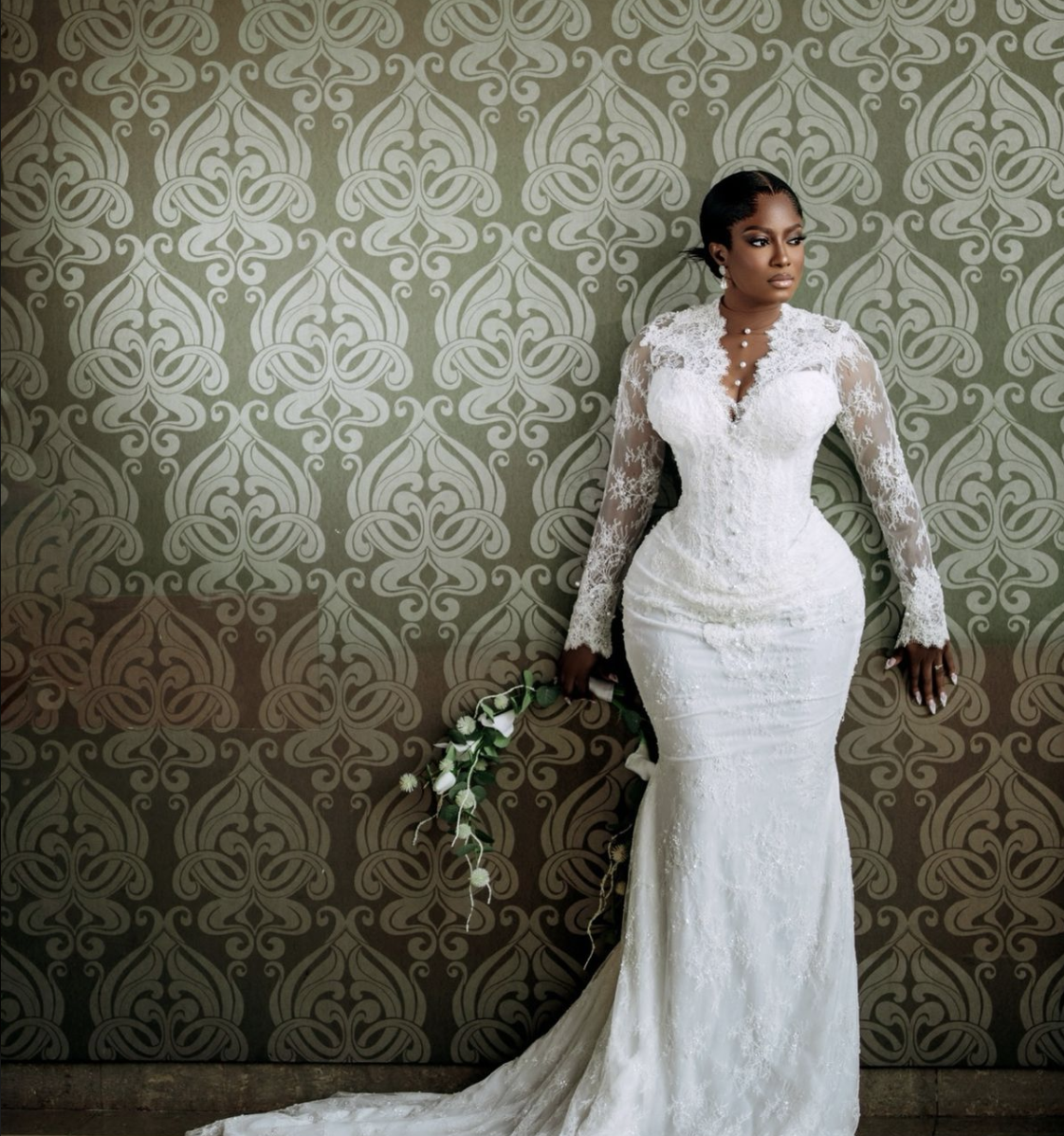 Bride in a white lace wedding gown holding a flower lei, standing against a decorative wall.