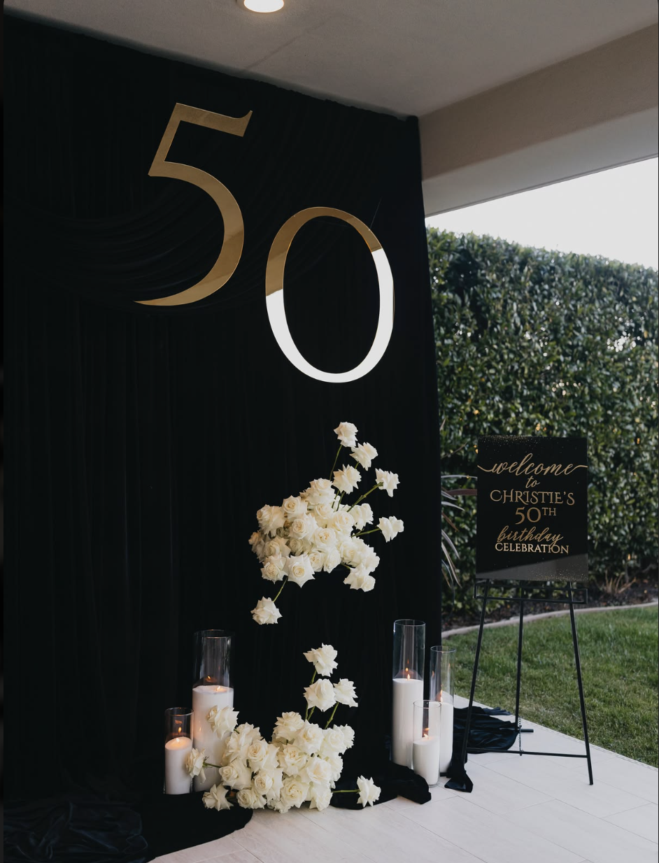Decorated entrance for Christie's 50th birthday celebration with large gold number 50, white floral arrangement, candles, and a sign welcoming guests.