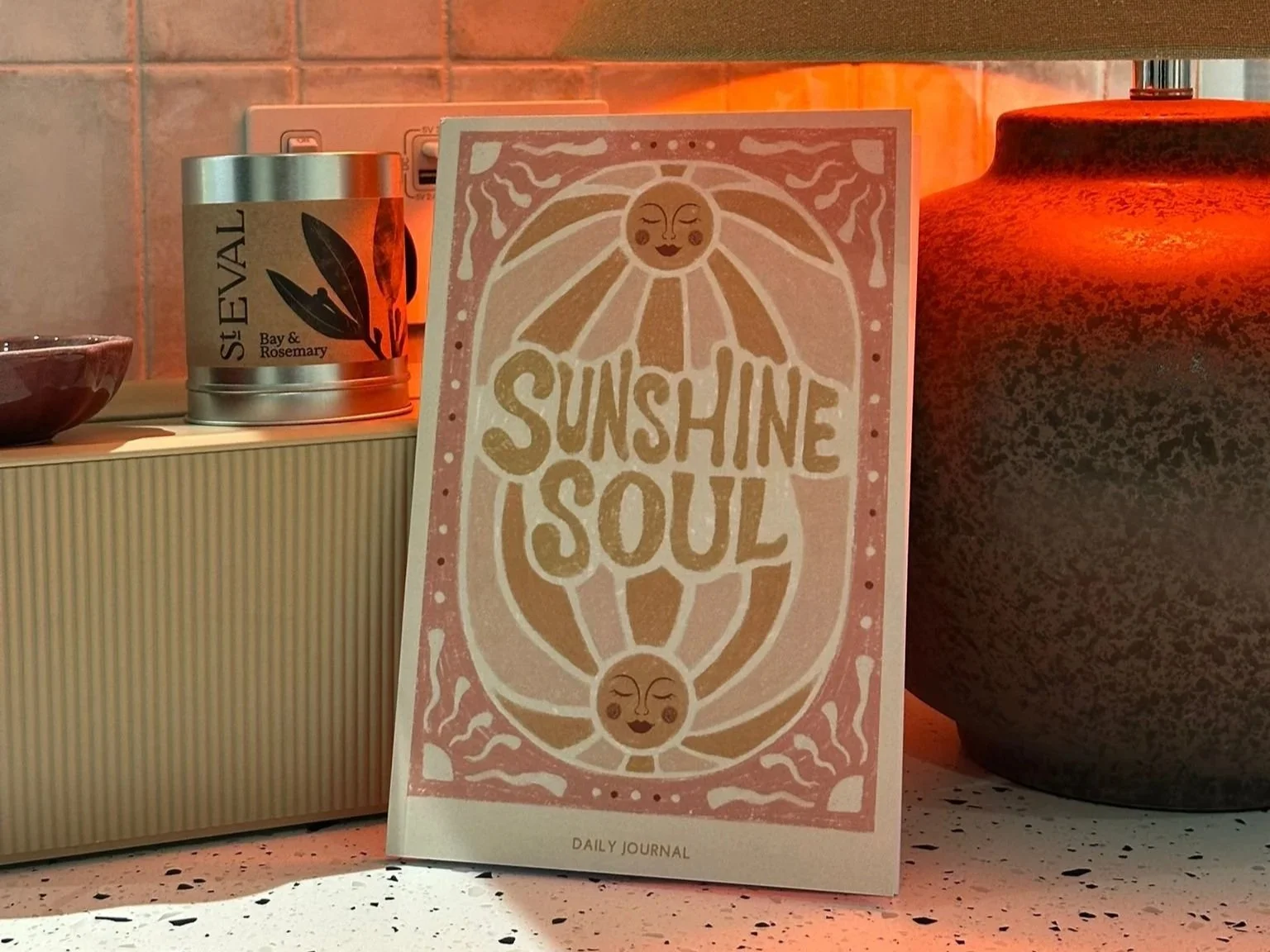 A daily journal with the phrase "Sunshine Soul" and a smiling sun illustration, surrounded by warm lighting on a countertop.