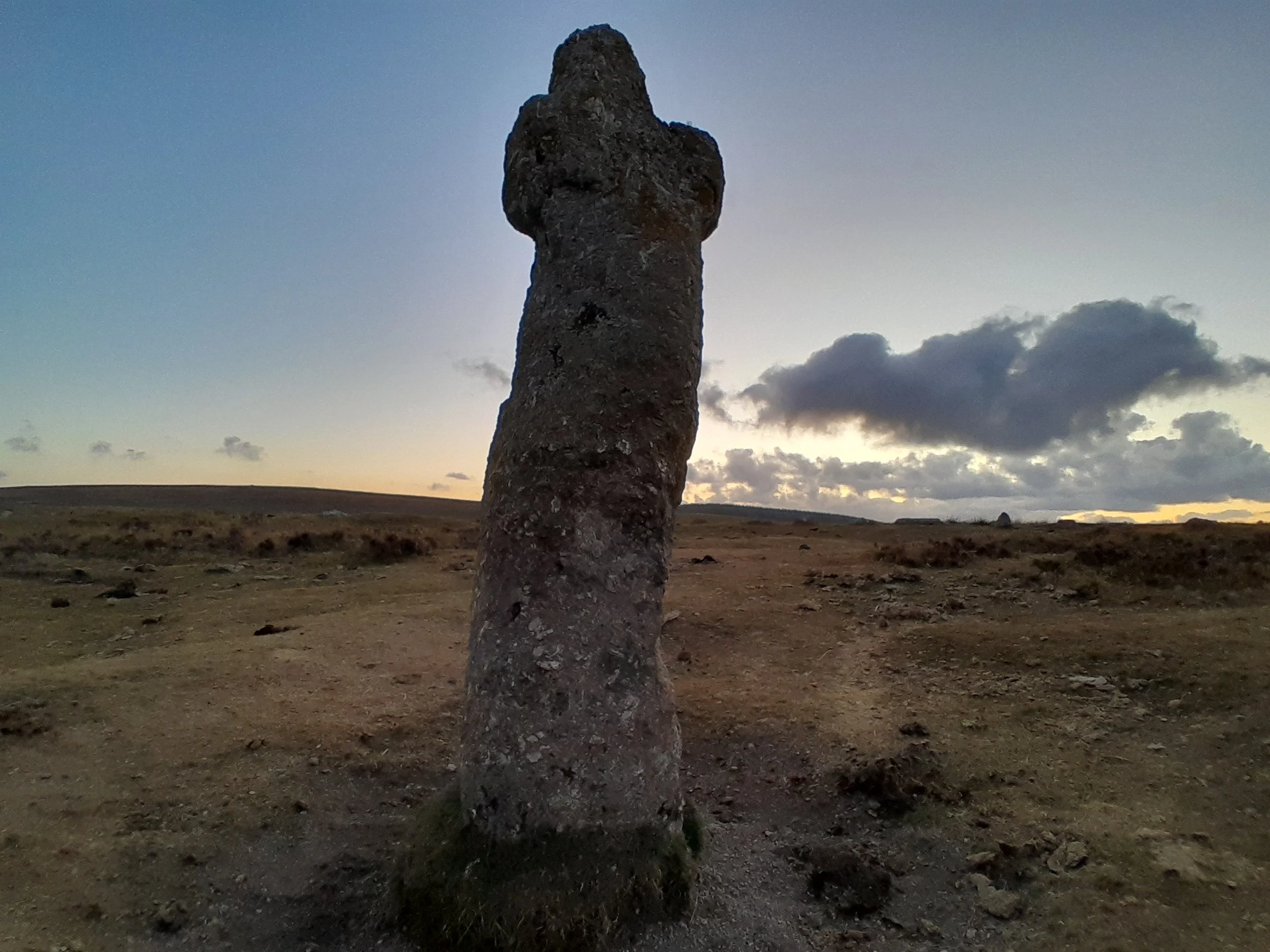 A lone standing stone in a vast open landscape during twilight with clouds in the sky.