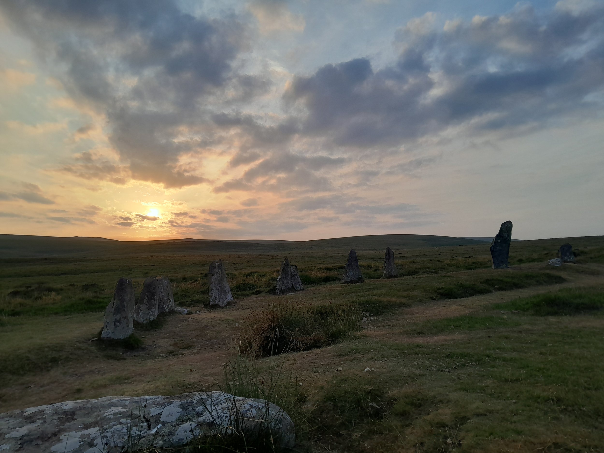A landscape at sunset with a cloudy sky, open grassland, and standing stones in the foreground.