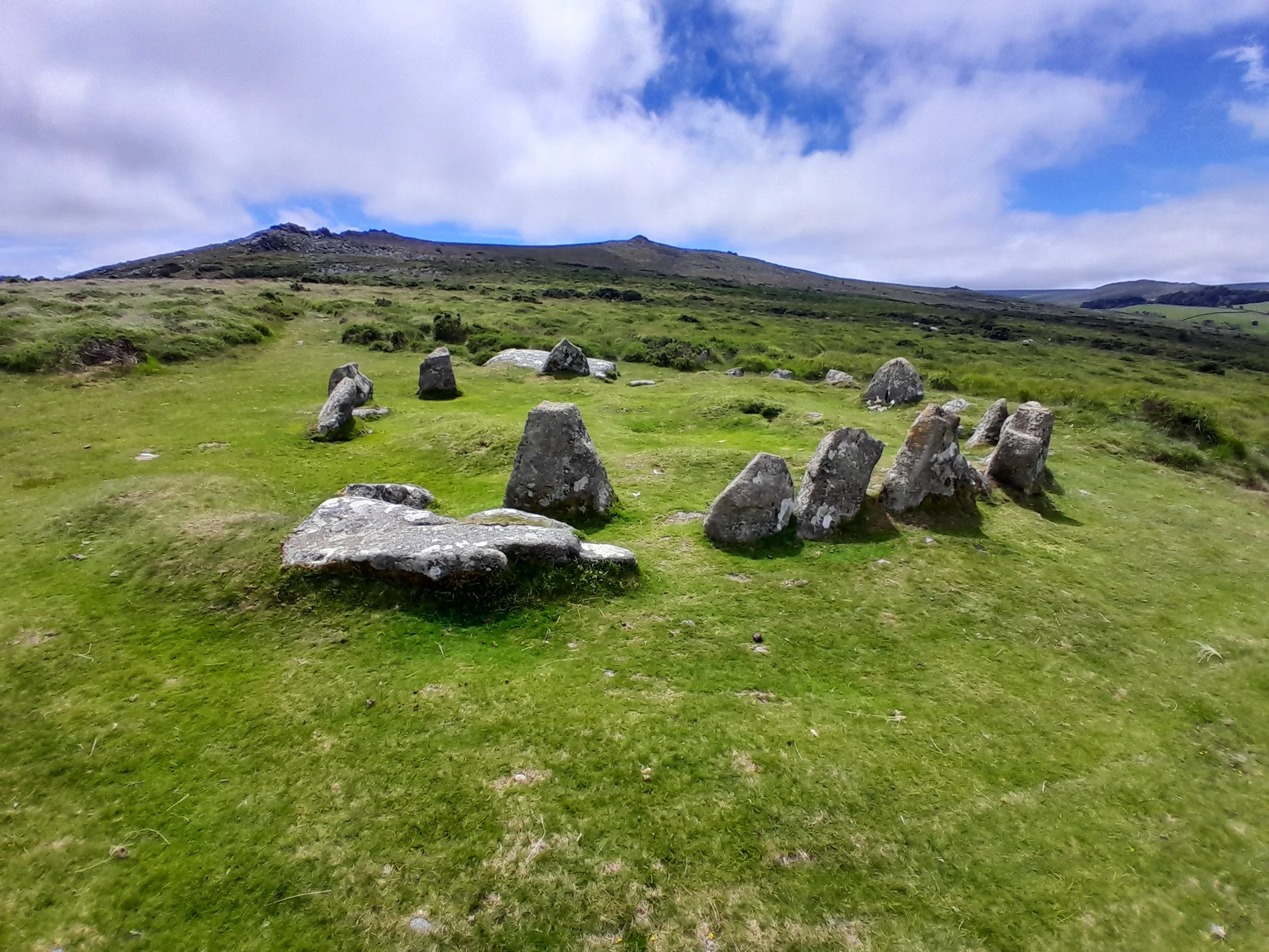 A grassy landscape with scattered rocks in the foreground, a hill in the background, and a mostly cloudy sky with patches of blue.