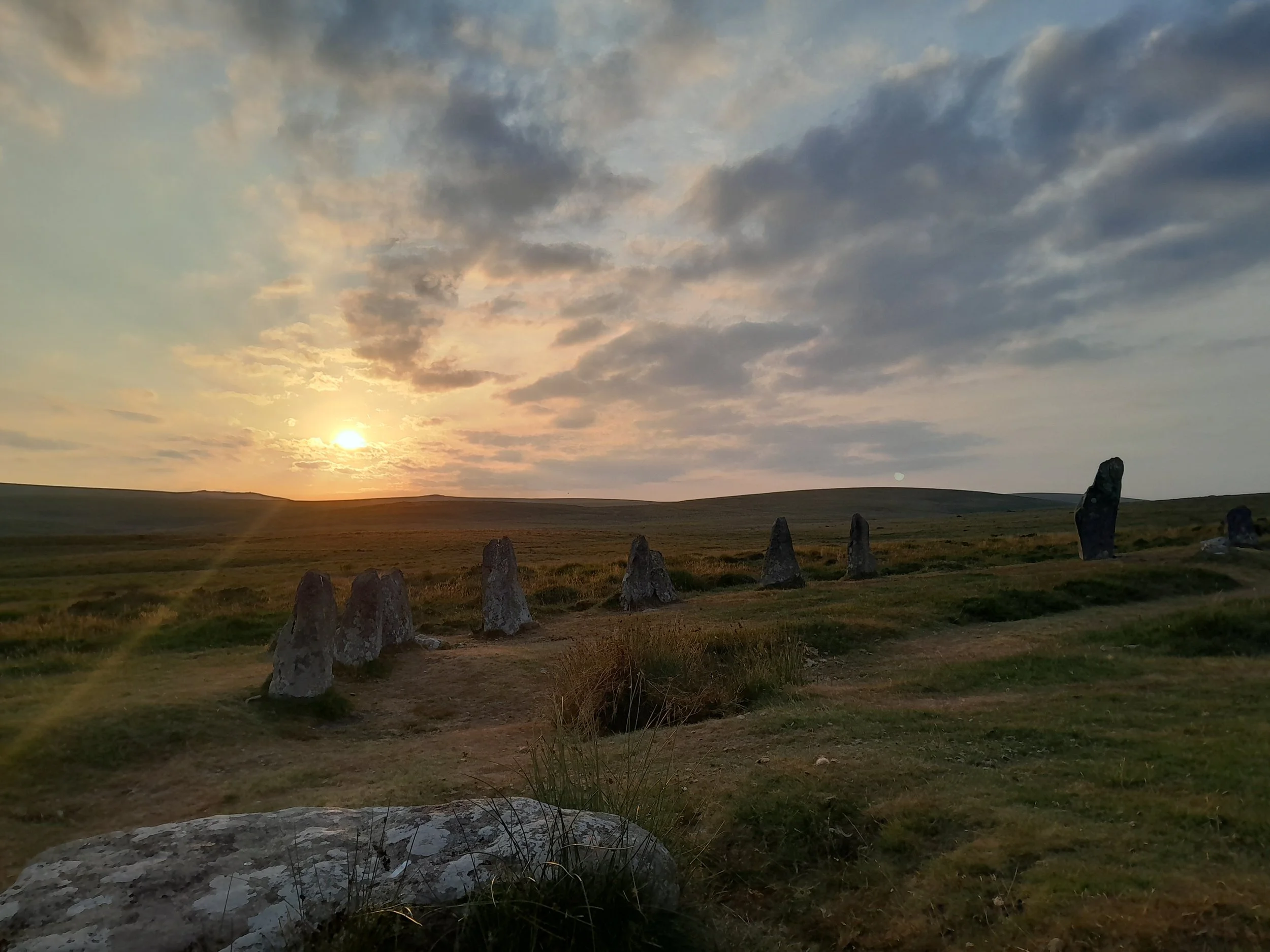 A landscape with grassy fields, standing stones, a sunset sky with clouds, and a hill in the distance.