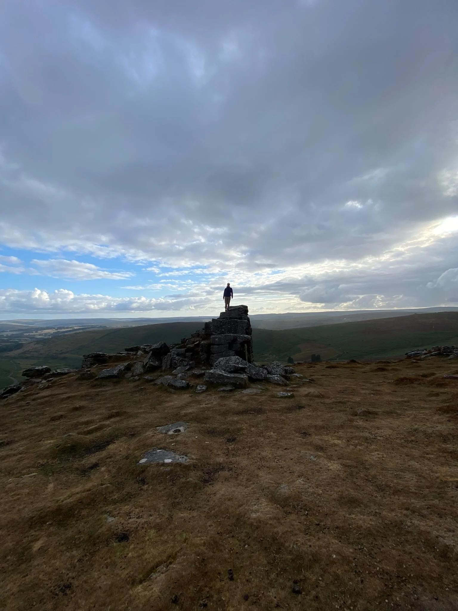 A person standing on a rocky formation in a wide open landscape with rolling hills in the distance and a cloudy sky overhead.