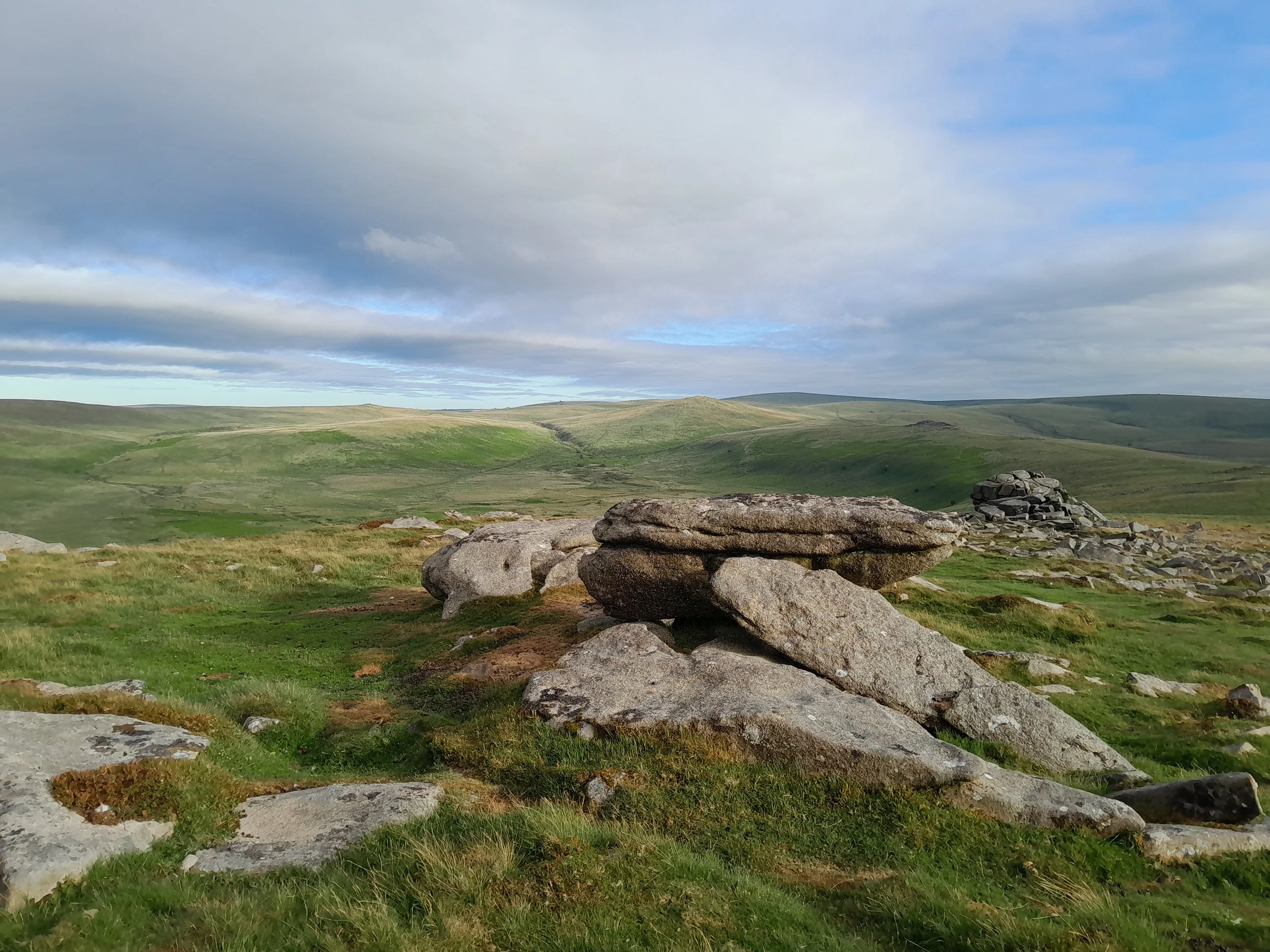 Open grassy landscape with rolling hills, large rocks in the foreground, and a partly cloudy sky overhead.