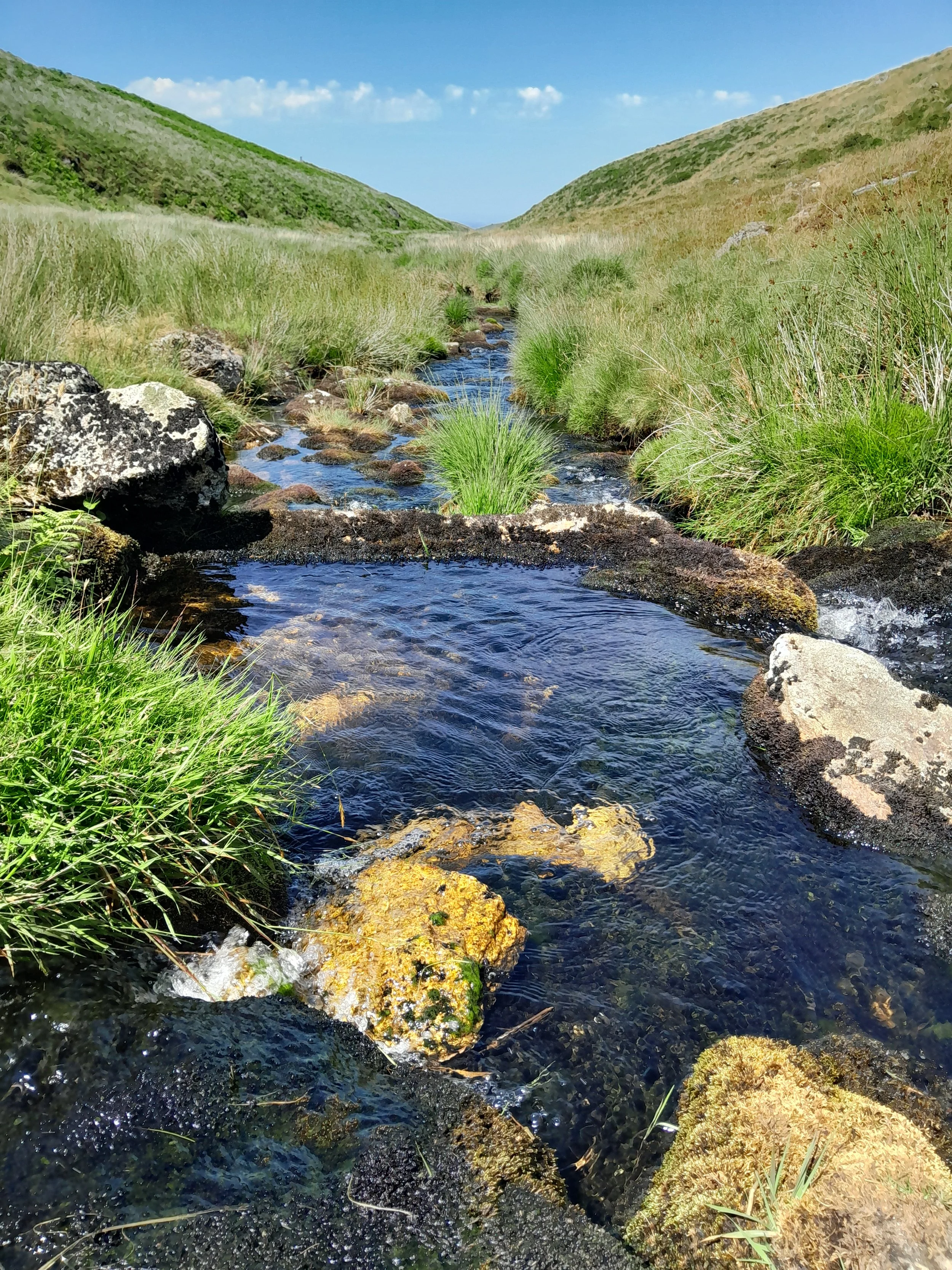A small stream flowing between grassy hills under a blue sky with white clouds.