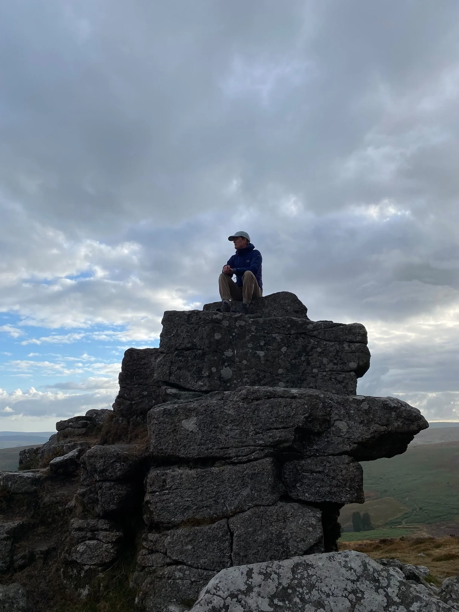 A person sitting on a large rock formation outdoors against a cloudy sky, with rolling landscape in the background.