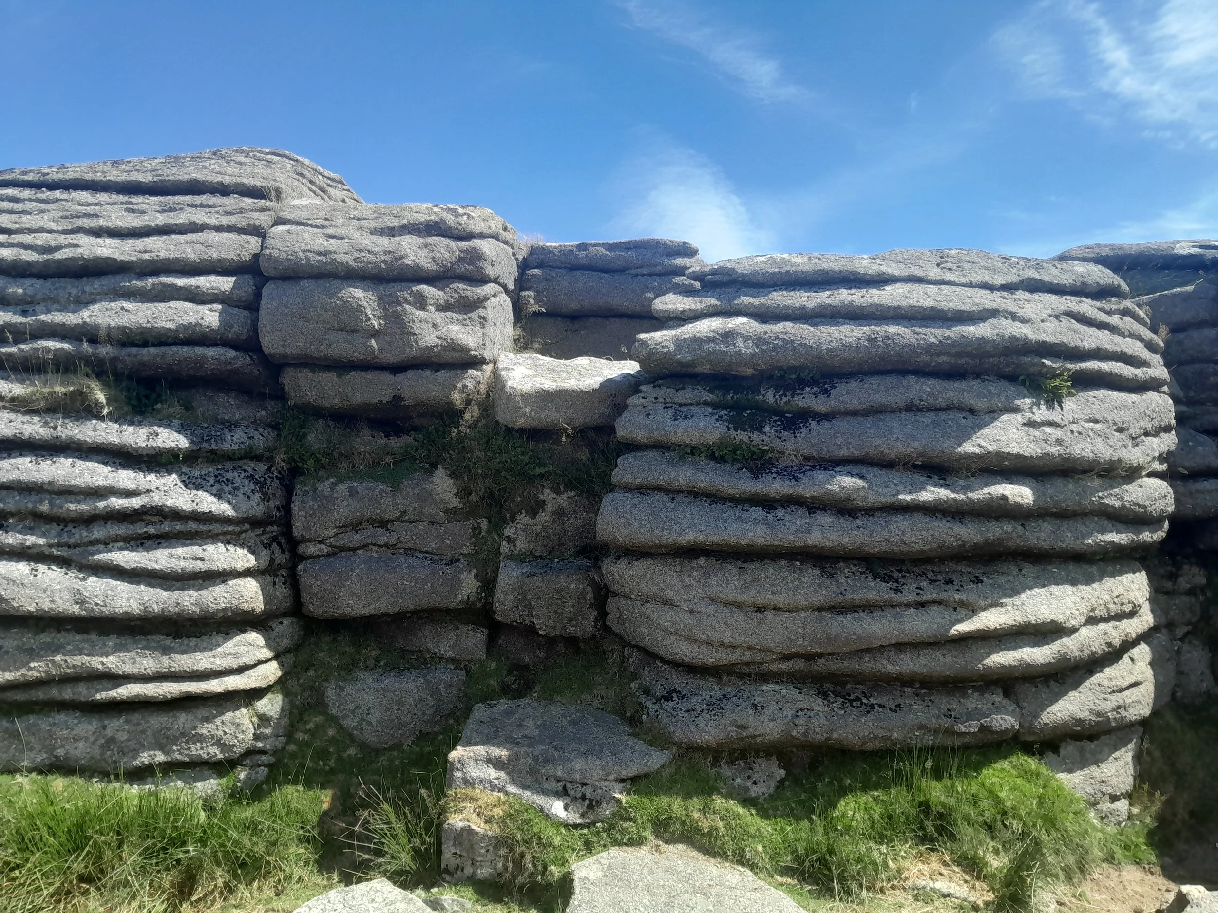 A dry-stacked stone wall with horizontal layers of large, flat rocks, with some small patches of grass and greenery at the base beneath a blue sky with wispy clouds.