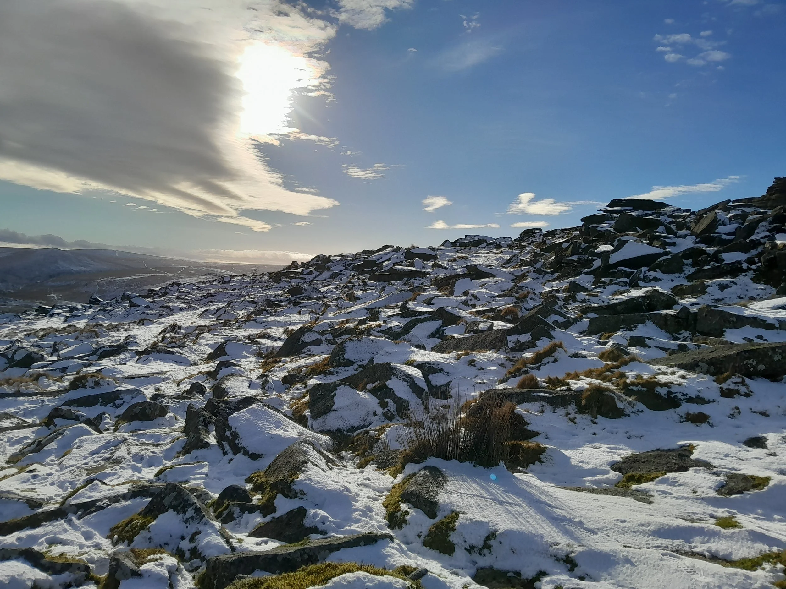 Snow-covered rocky hillside with patches of snow and grass, cloudy sky with sunlight peeking through, distant mountains and hills visible in the background.