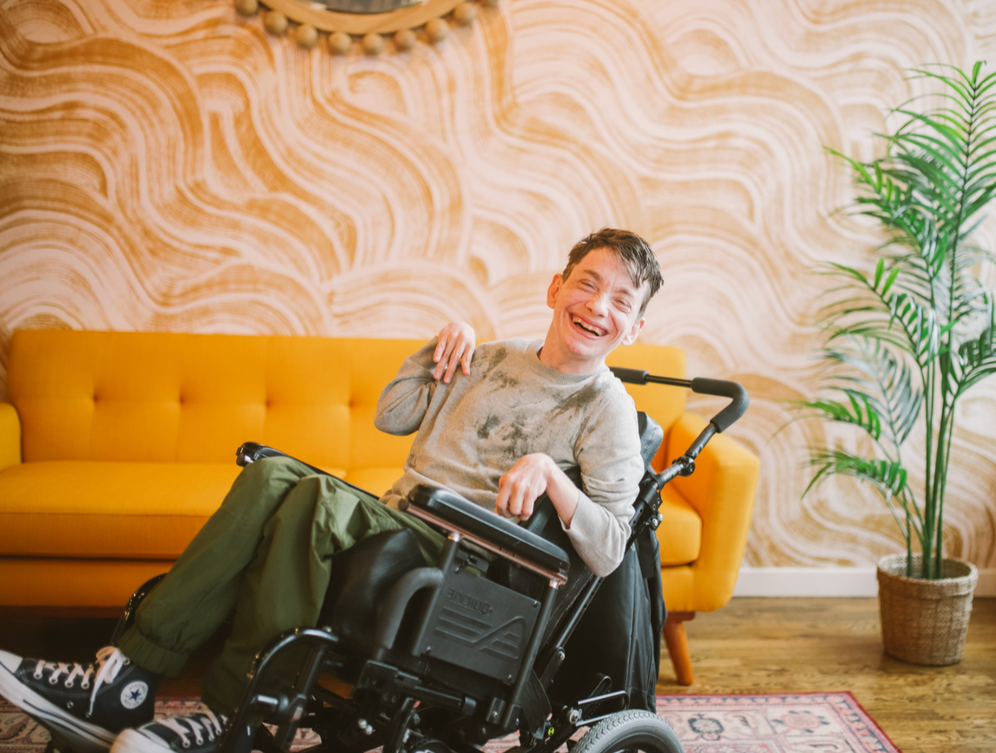 A young man in a wheelchair, smiling and laughing, in a room with a yellow sofa, patterned wall, and a potted plant.
