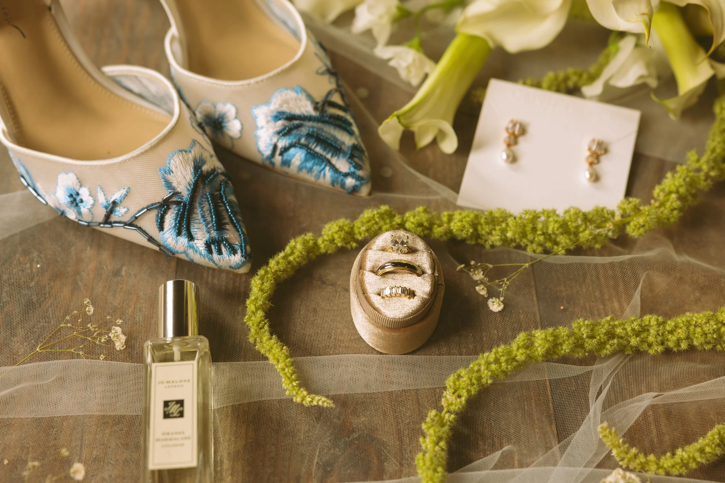 Close-up of bridal accessories including embroidered wedding shoes, pearl earrings, wedding rings, perfume, and floral decorations on a soft fabric background.