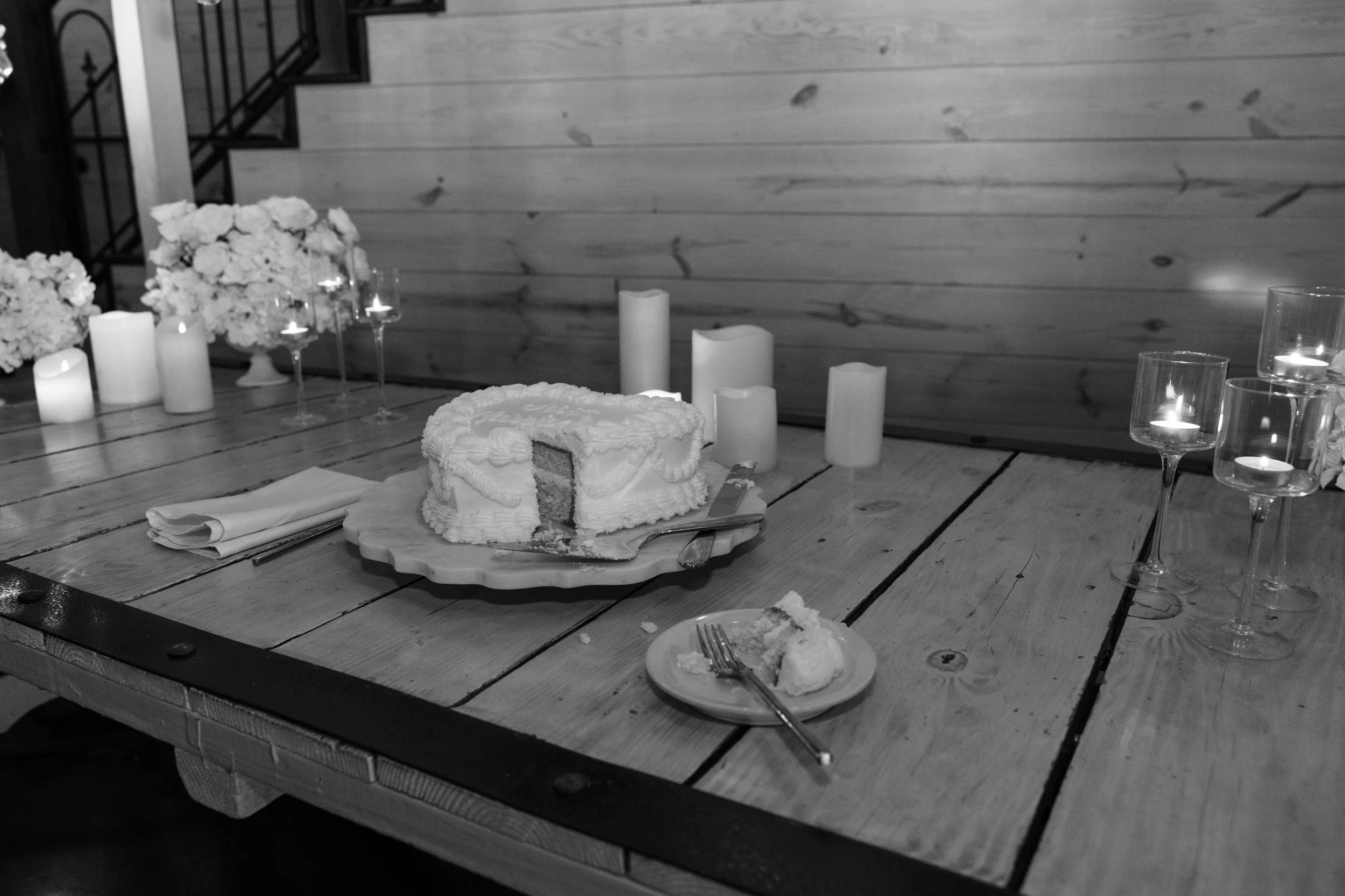 A partially eaten sheet cake with white icing on a wooden table, surrounded by candles, flowers, and empty dessert dishes with utensils, in a rustic setting.