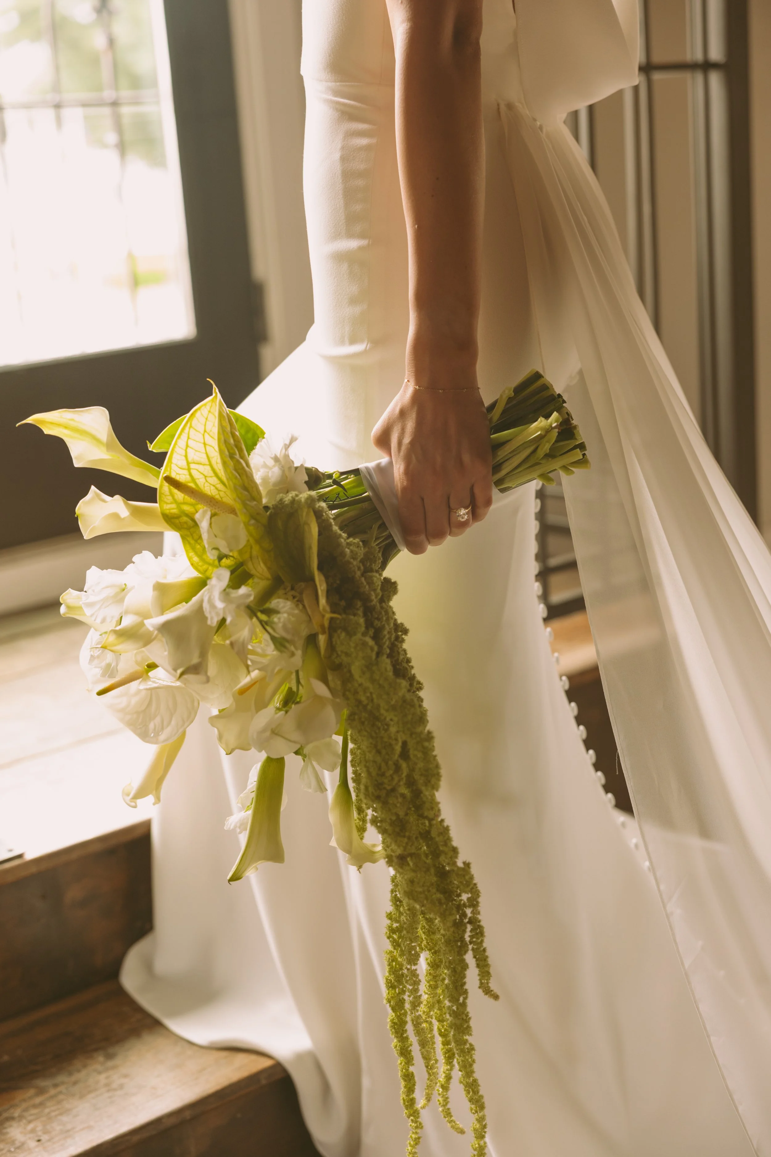 A woman in a wedding dress holding a bouquet of white and green flowers, standing near a window with sunlight.