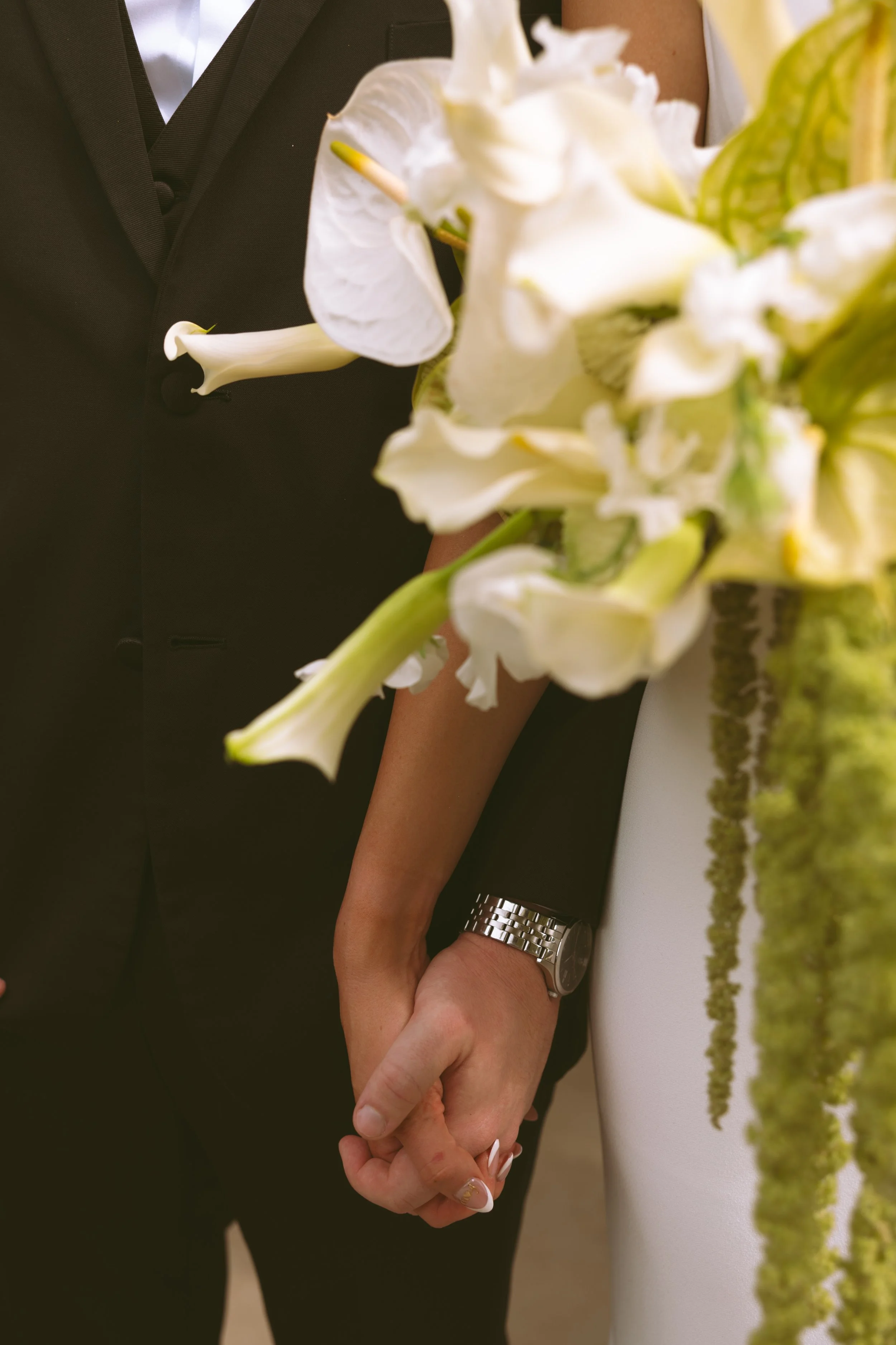 A close-up of a couple holding hands, dressed in formal attire, standing next to a white wedding dress and a bouquet of white flowers. The man is wearing a black suit, and the woman has a silver watch on her wrist.