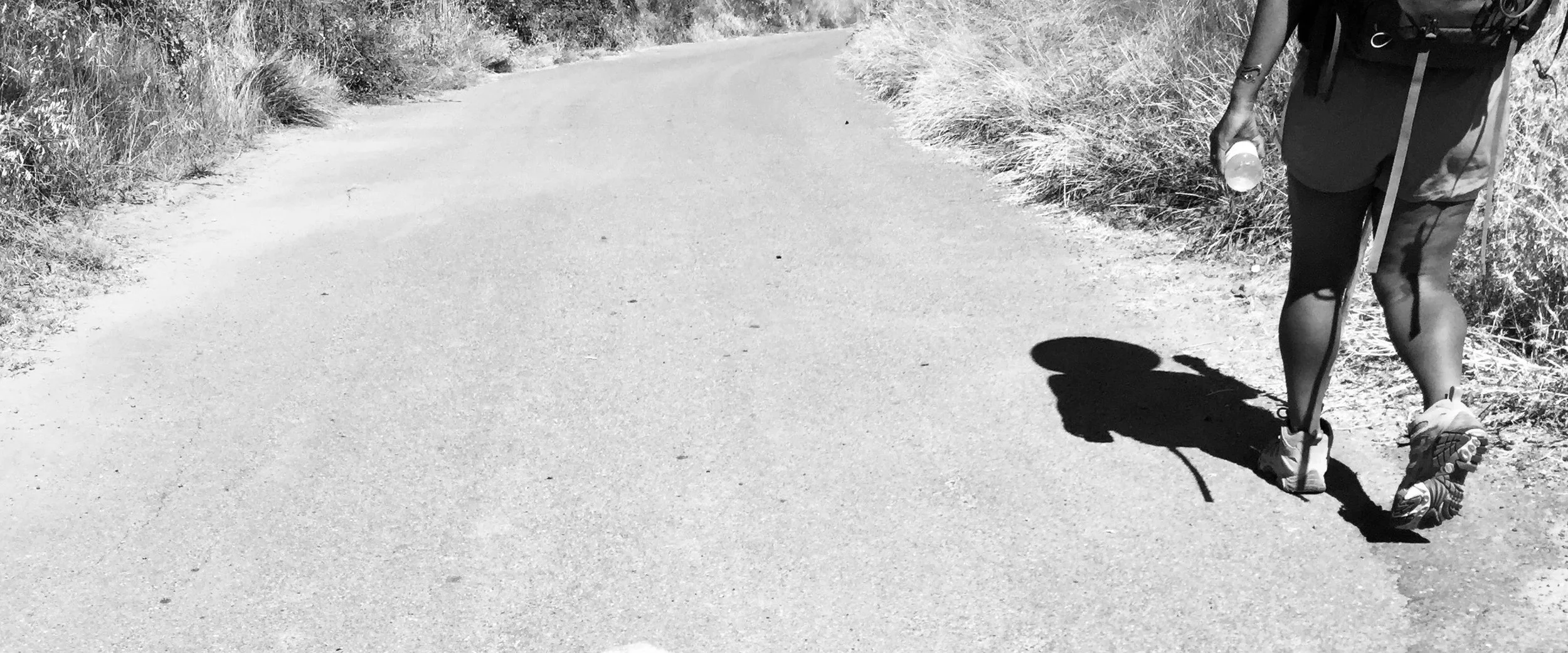 Person walking on a paved trail surrounded by bushes, holding a water bottle, with a prosthetic leg visible, in black and white.