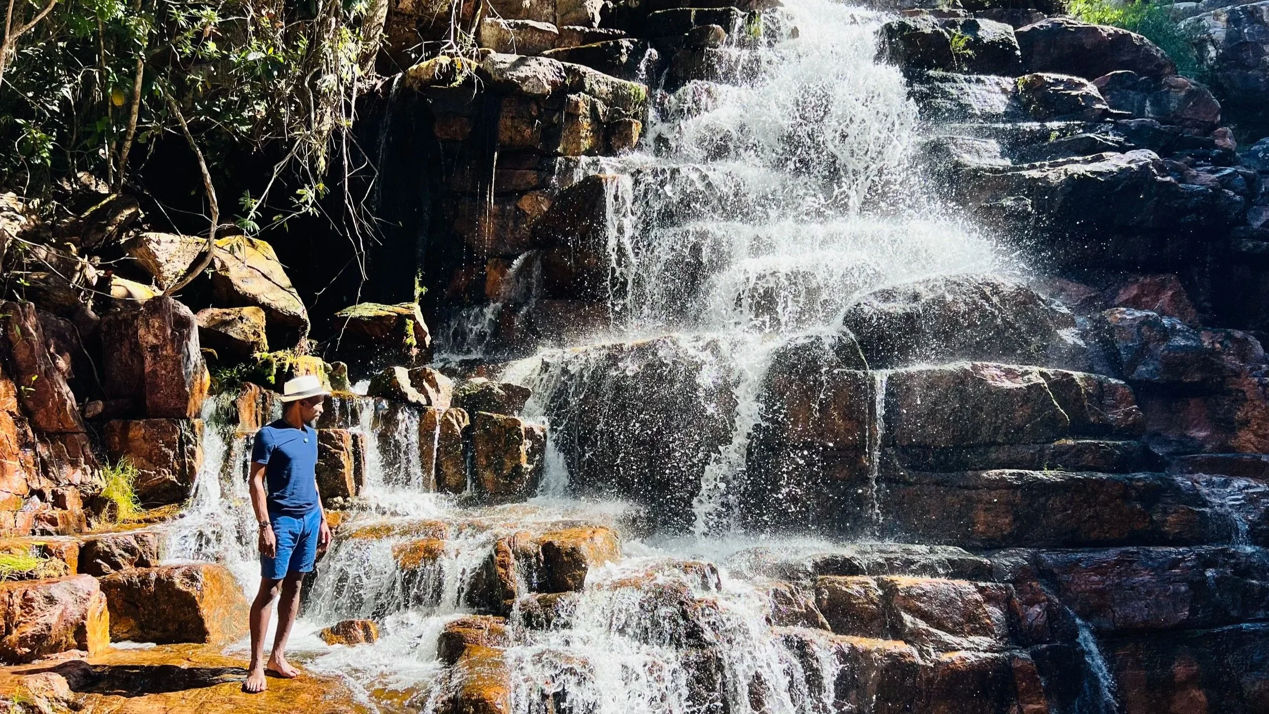 A person standing on rocks at the base of a waterfall with water cascading down large rocks surrounded by greenery.