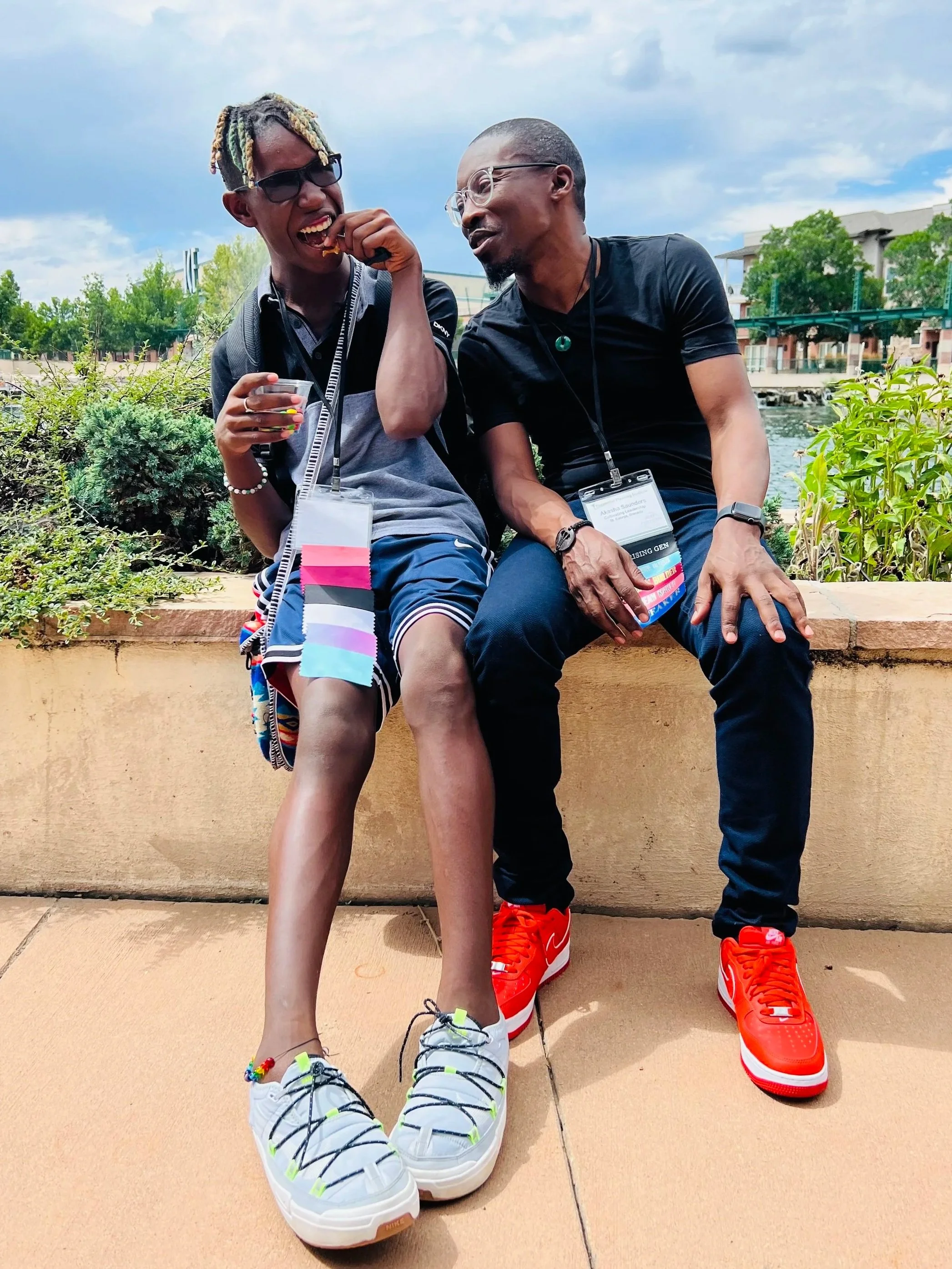 Two men sitting on a low wall outdoors by a waterway, wearing large shoes and shirtless, smiling and talking. One man is wearing a black shirt and glasses, the other is wearing a black shirt and glasses, with a lanyard and badge. Green trees and buildings are in the background.