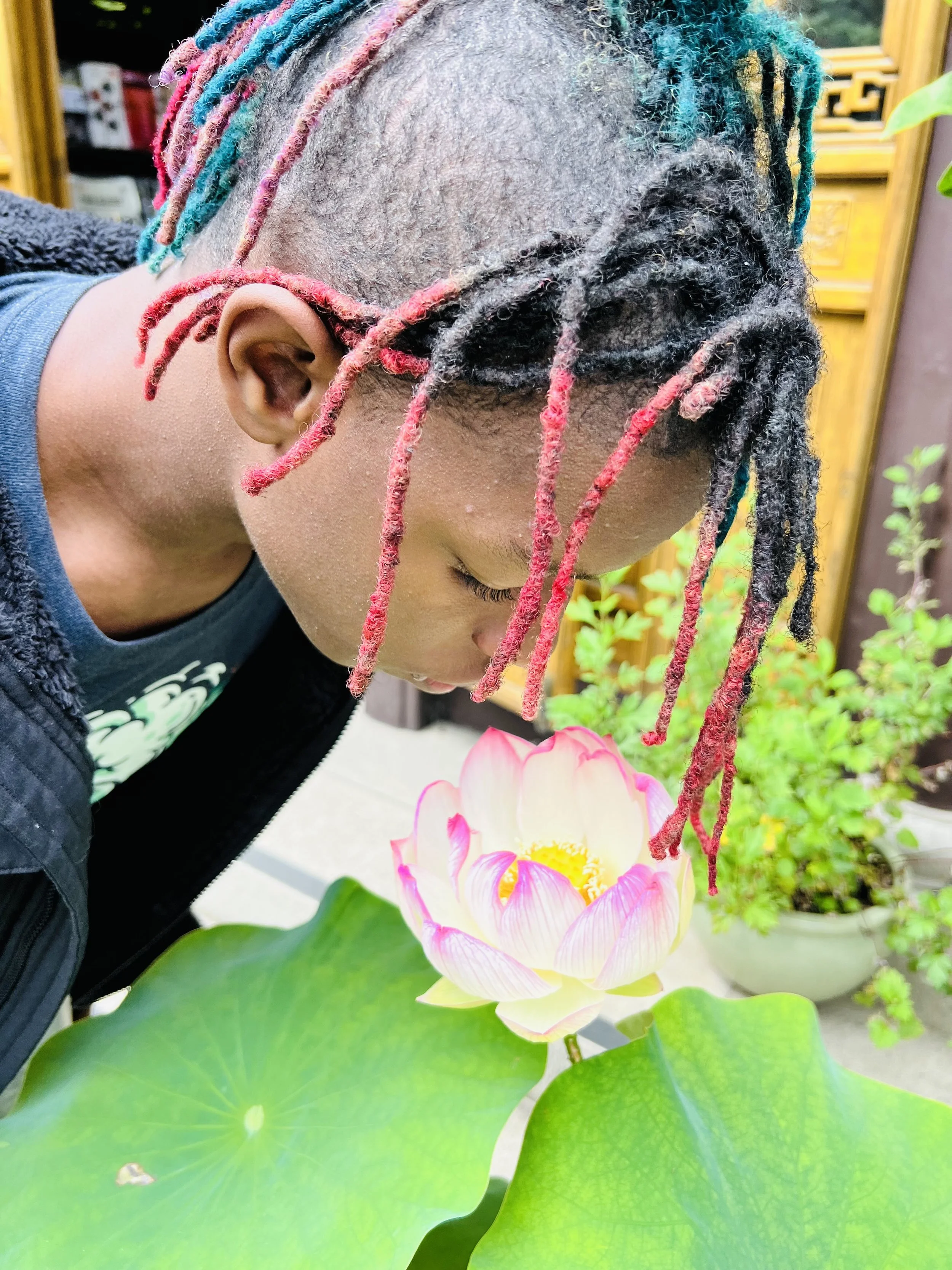 A person with colorful dreadlocks and a buzz cut hairstyle smelling a pink and white lotus flower. The person wears a dark jacket and the background includes greenery and a yellow wooden structure.