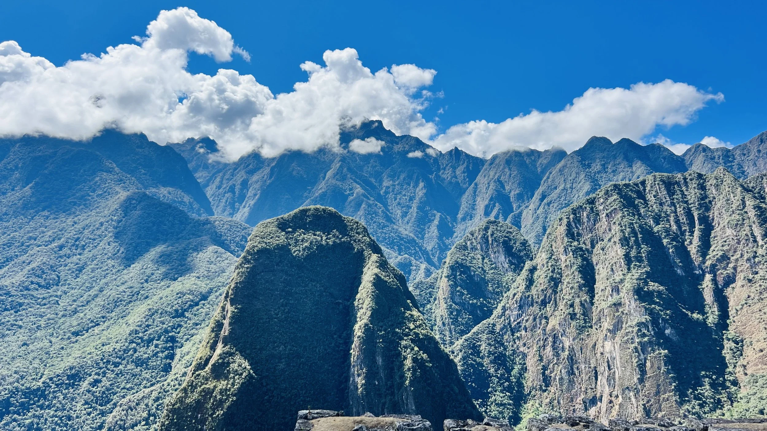 A panoramic view of lush green mountain peaks with a bright blue sky and white clouds overhead.