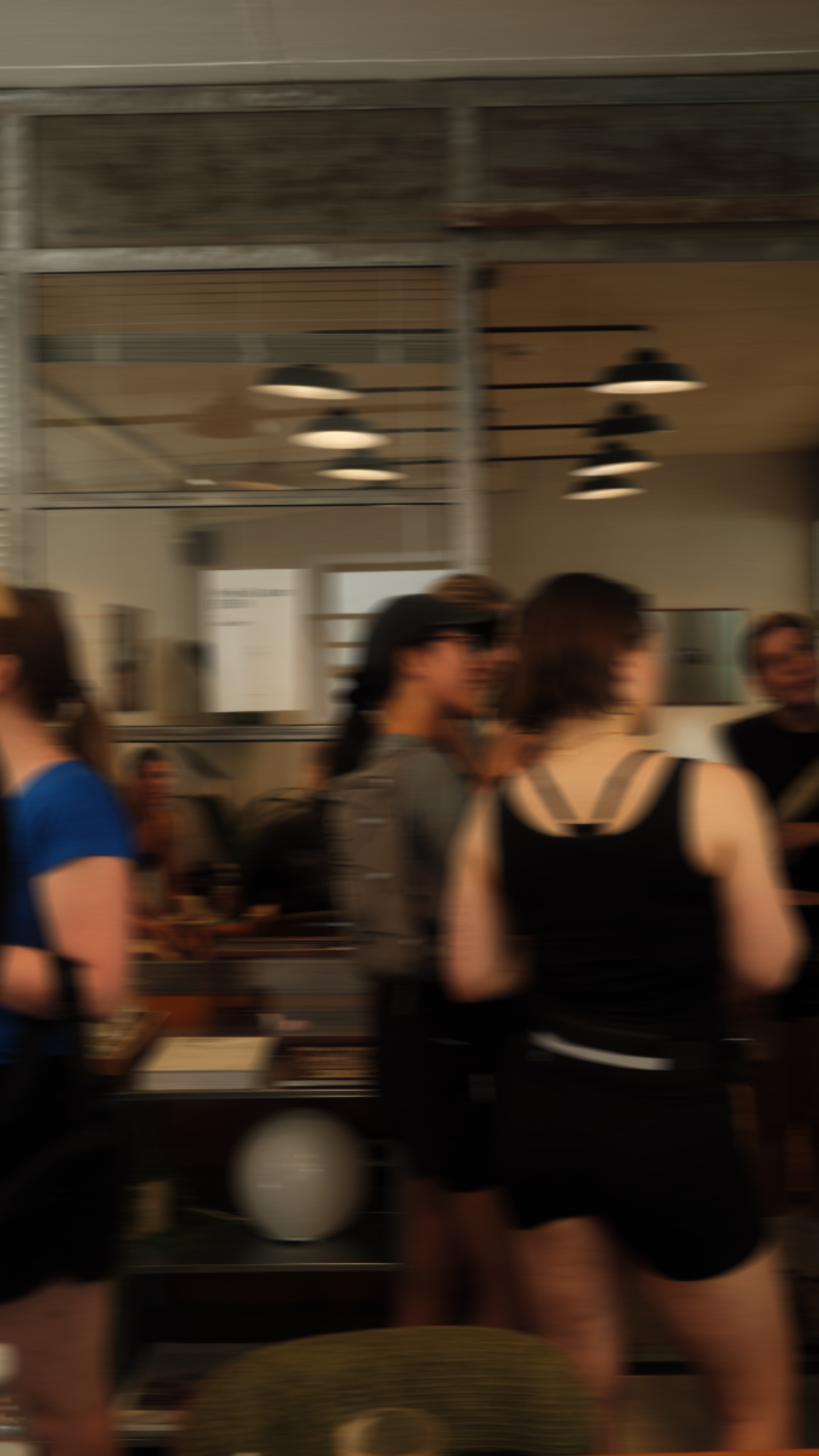 Menschen in einer modernen Cafeteria, einige Frauen, die sich unterhalten, mit Lampen an der Decke und einem offenen Küchenbereich im Hintergrund.