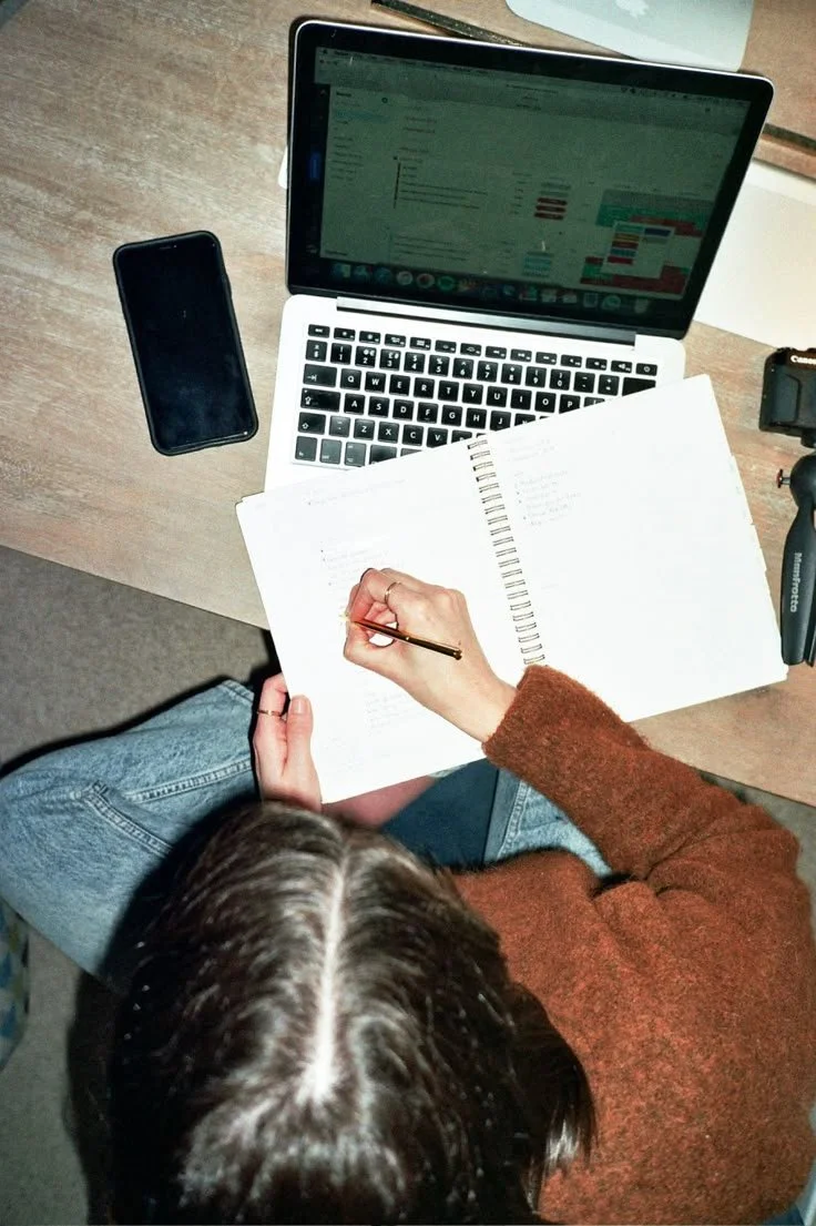 Eine Person schreibt in ein Notizbuch, während sie an einem Schreibtisch sitzt, mit einem Laptop und einem Smartphone vor sich.