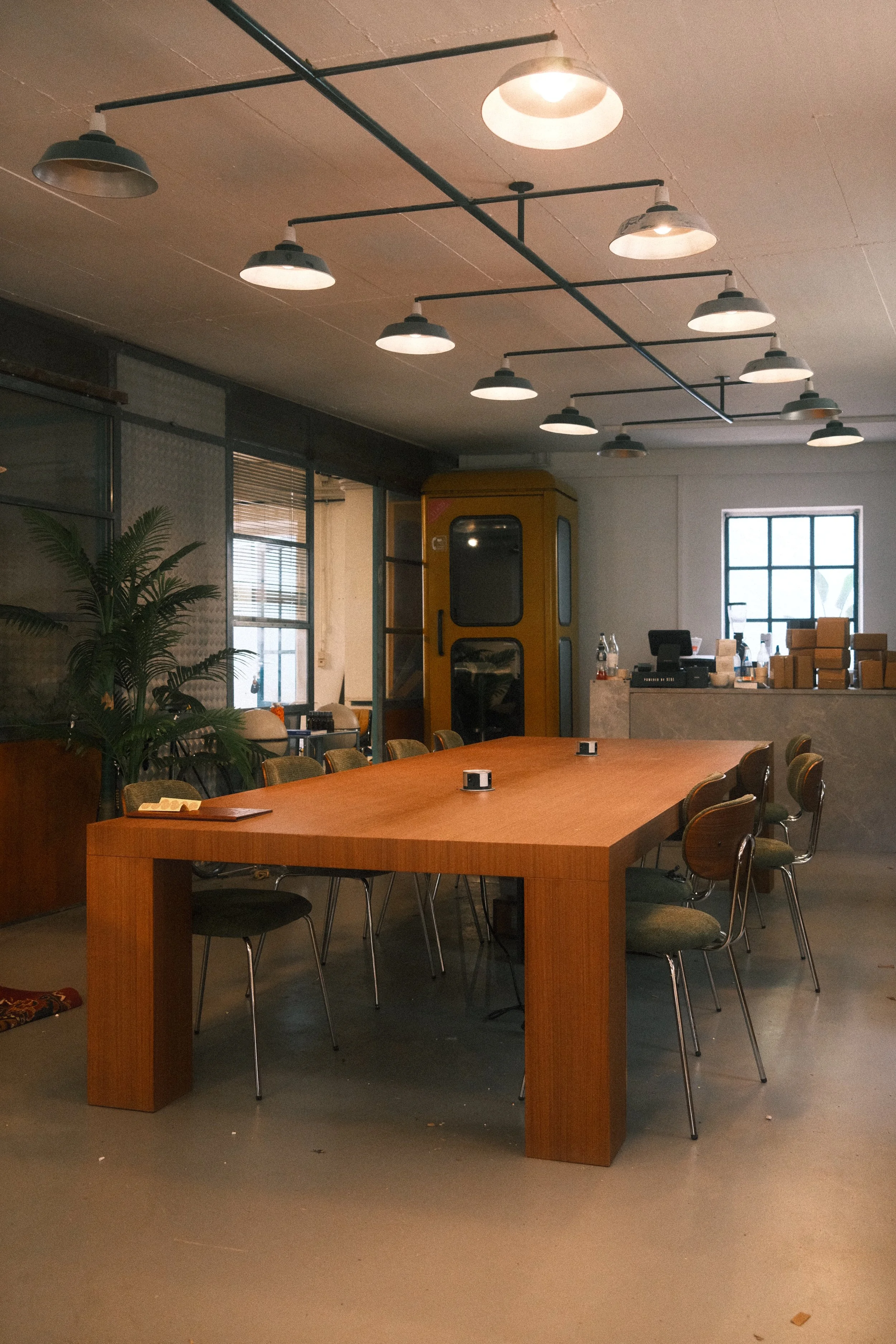 Innenansicht eines modernen Cafés oder Restaurants mit großem Holztisch, mehreren Stühlen, Pflanzen, großen Fenstern und industriellen Deckenleuchten.