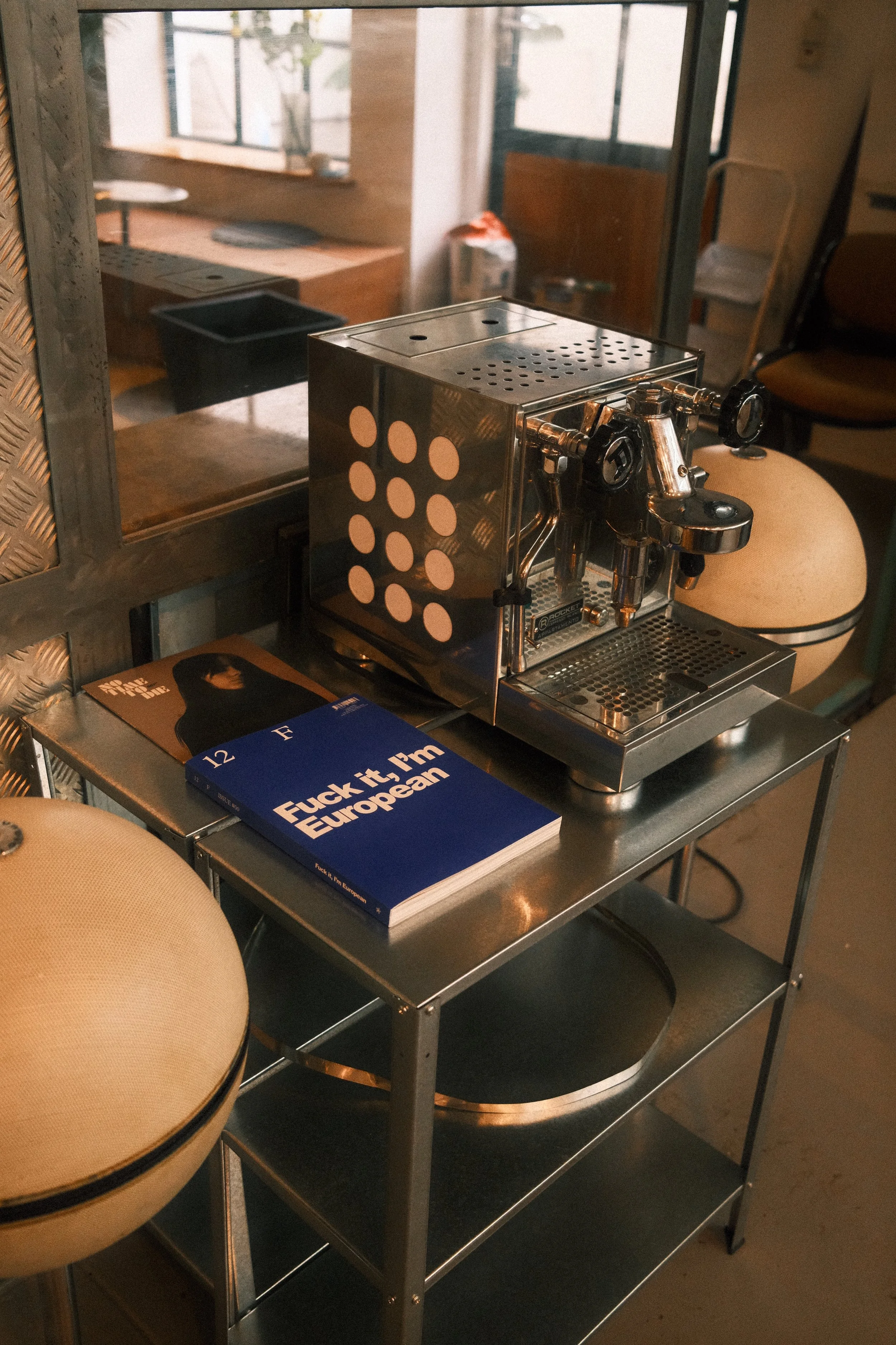 Ein Espressogerät auf einem Metallwagen neben einem beige Stuhl, auf dem ein Buch liegt.