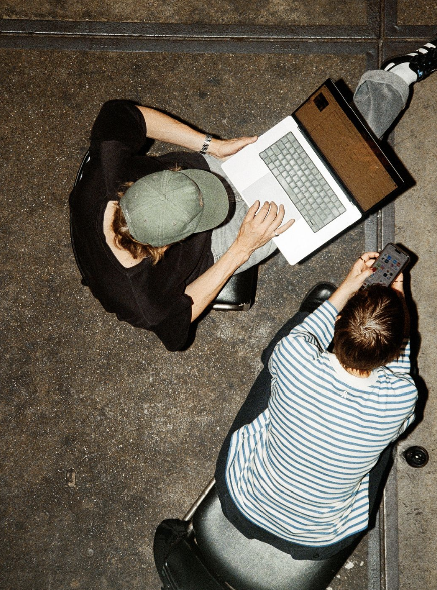 Zwei Personen sitzen auf dem Boden, eine mit einem Laptop und die andere mit einem Smartphone, auf einem dunklen Bodenbelag. Eine Person trägt ein grünes Cap, die andere einen gestreiften Pullover.